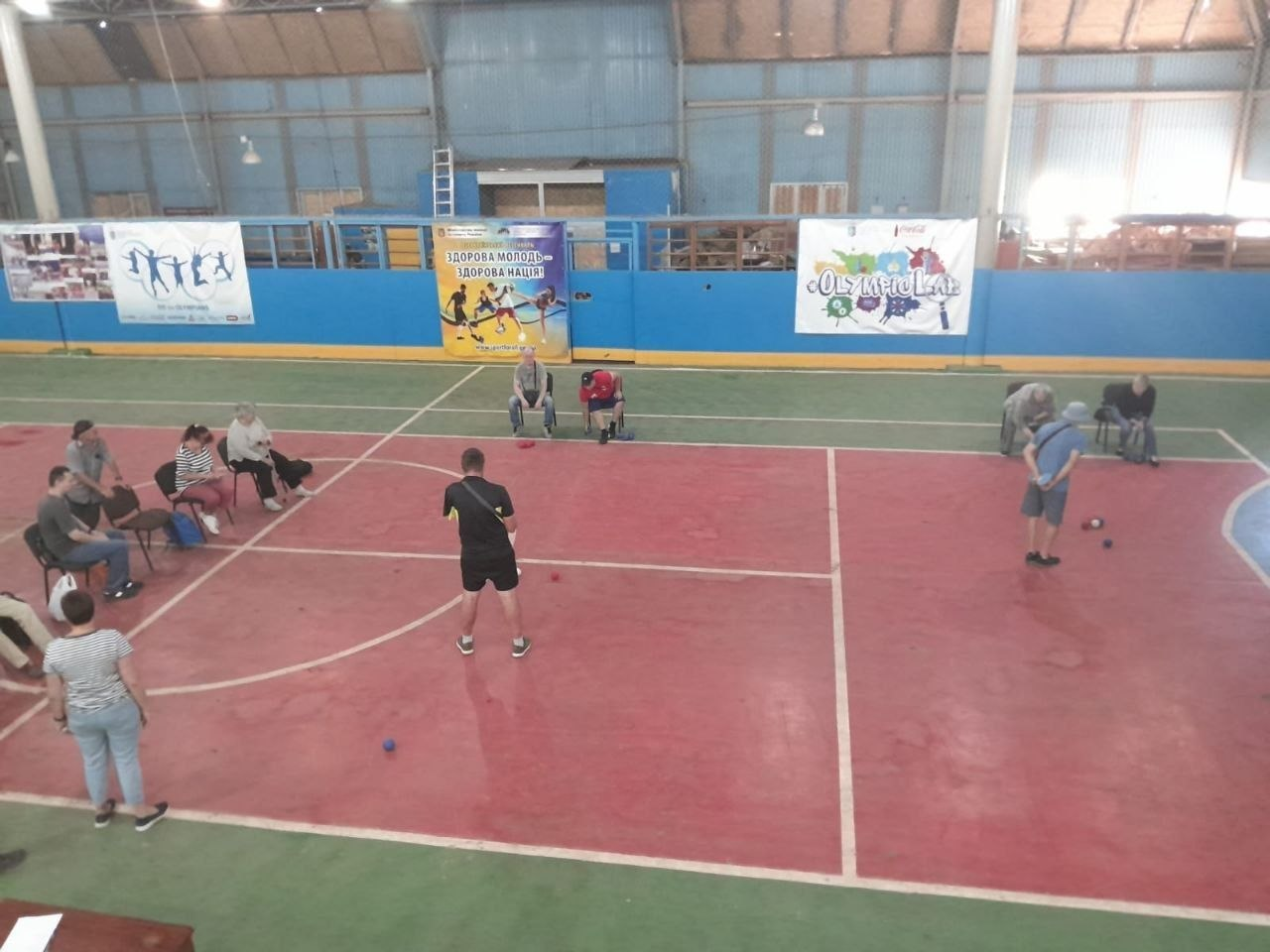 Спортсмени під час гри на спеціально розміченому червоно-зеленому майданчику у критому спортзалі. Кілька учасників зосереджено кидають кулі на дальність та точність, намагаючись поцілити ближче до цілі, тоді як інші гравці та вболівальники спостерігають за процесом, сидячи на стільцях уздовж ліній поля. На синіх стінах зали розвішані спортивні банери з гаслами «Здорова молодь — здорова нація!» та олімпійською символікою. Фотографія ілюструє ігровий процес змагань із боча серед людей з порушеннями слуху, які відбулися на Херсонщині до Дня фізичної культури і спорту України, демонструючи наполегливість учасників та розвиток адаптивного спорту.