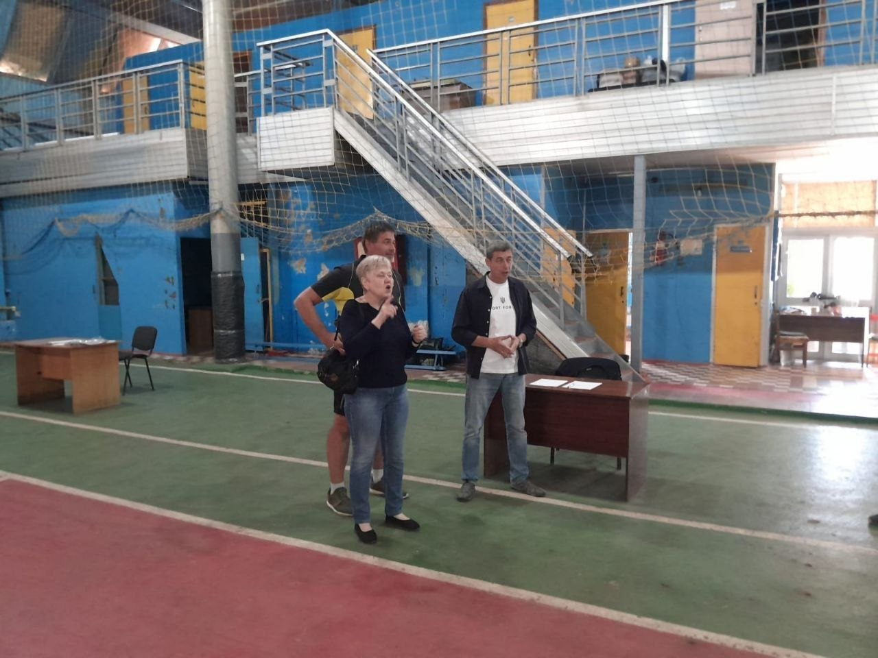 Організатори та судді змагань стоять на зеленому покритті ігрового майданчика у критому спортзалі поруч із суддівським столом та виступають перед учасниками. Жінка на передньому плані емоційно спілкується за допомогою жестів, здійснюючи сурдопереклад або жестовий супровід для спортсменів. Позаду неї стоять двоє чоловіків у спортивному та повсякденному одязі, які допомагають у проведенні заходу. На задньому плані видно сині стіни зали, захисну сітку та металеві сходи на другий ярус будівлі. Фотографія ілюструє організаційну частину змагань із боча серед людей з порушеннями слуху, які відбулися на Херсонщині до Дня фізичної культури і спорту України, забезпечуючи безбар'єрність, доступність інформації та комфортні умови для всіх учасників турніру.