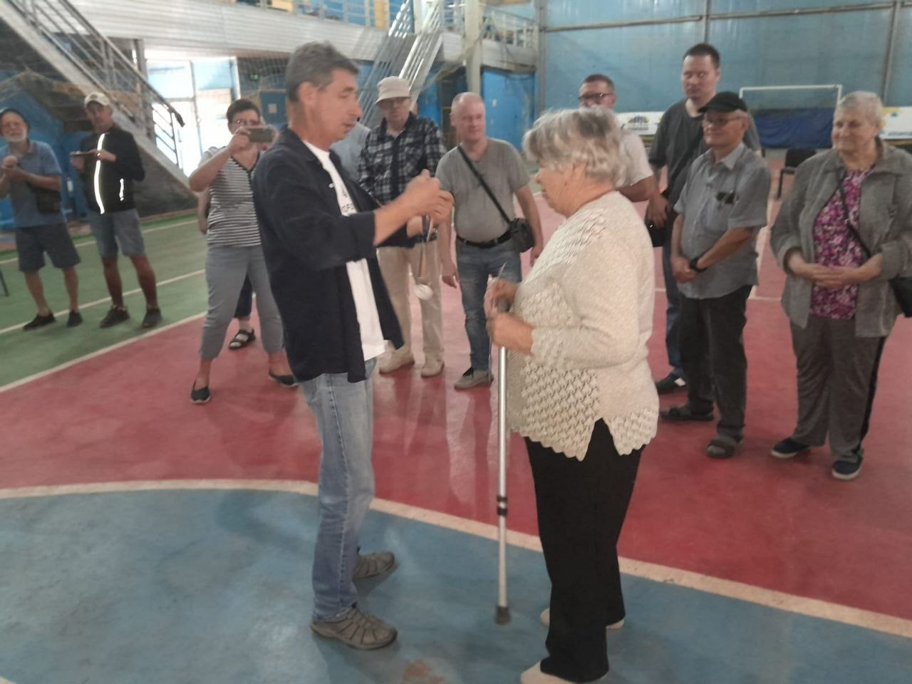Урочистий момент нагородження переможців змагань у критому спортзалі з червоно-синім покриттям підлоги. Чоловік у синій сорочці та джинсах одягає нагородну медаль на шию літній жінці у світлій кофті, яка спирається на тростину. Інші учасники заходу та вболівальники стоять поруч і радісно спостерігають за процесом, а жінка ліворуч фотографує подію на смартфон. Фотографія ілюструє церемонію нагородження призерів змагань із боча серед людей з порушеннями слуху, які відбулися на Херсонщині до Дня фізичної культури і спорту України, підкреслюючи жагу до перемоги, визнання здобутків та підтримку активного довголіття.