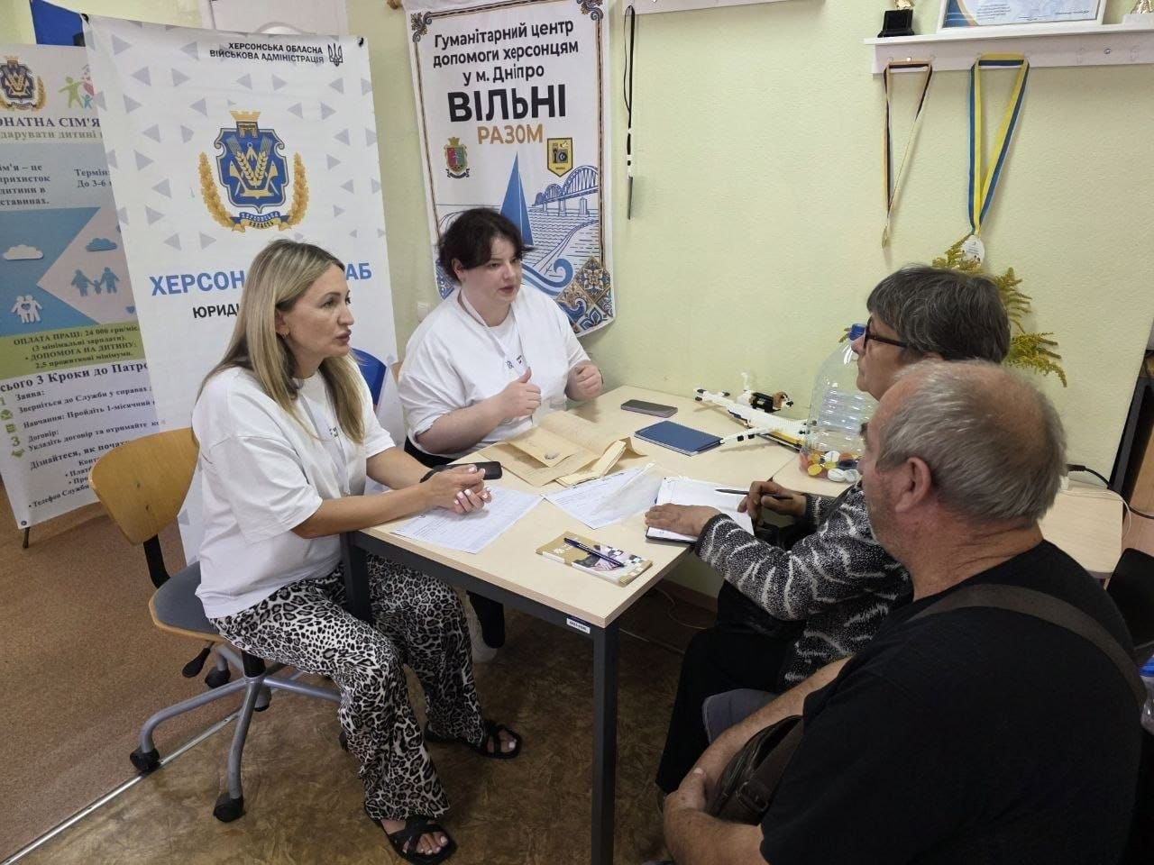 Дві жінки-консультантки сидять за робочим столом у кабінеті хабу та надають соціальну допомогу відвідувачам — чоловіку та жінці похилого віку. На столі розкладені анкети, паперові конверти, робочі документи та канцелярія. Зліва від столу розміщено великий інформаційний банер Херсонської обласної військової адміністрації з написом Херсонський хаб. Юридична допомога, а на стіні позаду висить прапор із символікою під назвою Гуманітарний центр допомоги херсонцям у м. Дніпро «Вільні разом». Захід є частиною системної підтримки переселенців із Херсонщини в координаційному центрі, де кейс-менеджери благодійного фонду GLOBA22 та громадської організації Аваліст провели 8 консультацій щодо соціального супроводу з метою соціальної адаптації, вирішення побутових питань та підвищення життєстійкості внутрішньо переміщених осіб.