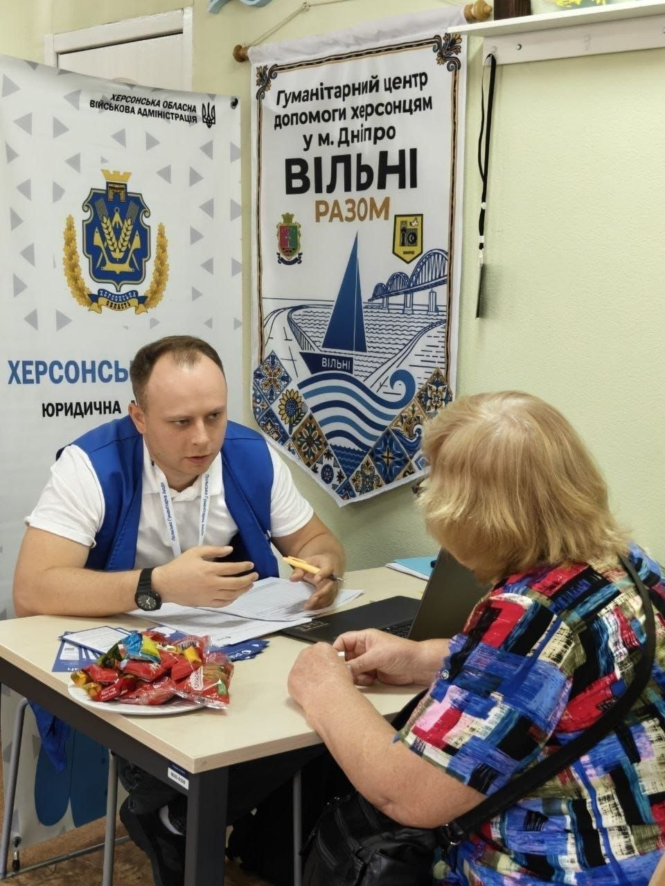 Чоловік-консультант у синьому брендованому жилеті сидить за робочим столом у кабінеті хабу та уважно розмовляє з жінкою старшого віку, яка сидить навпроти. Спеціаліст тримає в руці ручку й пояснює інформацію, спираючись на розкладені документи, а перед відвідувачкою на столі стоїть відкритий ноутбук та тарілка з солодощами. Ліворуч позаду чоловіка розміщено великий інформаційний банер Херсонської обласної військової адміністрації з написом Херсонський хаб. Юридична допомога, а на стіні висить фірмовий прапор із символікою Гуманітарний центр допомоги херсонцям у м. Дніпро «Вільні разом». Захід є частиною системної підтримки переселенців із Херсонщини в координаційному центрі, де фахівці благодійного фонду GLOBA22, громадської організації Аваліст, Міжнародного комітету порятунку (IRC) та Польської Гуманітарної Акції (PAH) надали 17 індивідуальних консультацій для правової та соціальної підтримки внутрішньо переміщених осіб.