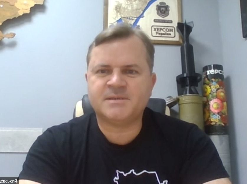 Скріншот робочої онлайн-наради за участі заступника начальника Херсонської ОВА Володимира Клюцевського, який сидить у кабінеті перед вебкамерою на тлі карти України, сувенірної таблички «ХЕРСОН Україна» та розписаних військових трофеїв. Візуальний матеріал ілюструє засідання Гуманітарного штабу, під час якого було затверджено розподіл допомоги (генераторів, палива, інструментів та питної води) для медичних закладів, комунальних підприємств та пунктів тимчасового проживання області.