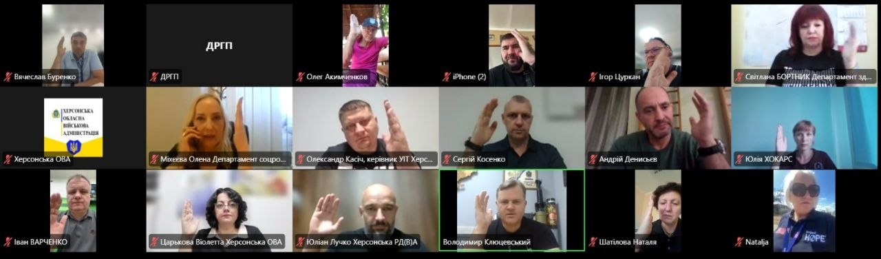 Скріншот онлайн-наради у форматі відеоконференції, яка ілюструє процес колективного ухвалення рішень під час чергового засідання Гуманітарного штабу при Херсонській ОВА. На екрані відображається сітка з вікон учасників, які тримають підняту руку під час голосування. У нижньому ряду по центру в зеленому рамковому вікні засідання проводить заступник начальника Херсонської ОВА Володимир Клюцевський. До обговорення долучилися заступник начальника Херсонської РВА Юліан Лучко, директорка Департаменту соціального розвитку Олена Міхєєва ХОДА, директорка Департаменту реалізації гуманітарної політики ХОДА Віолетта Царькова, начальник управління інформаційних технологій ХОДА Олександр Касіч, а також інші учасники наради, зокрема Вячеслав Буренко, Сергій Косенко, Андрій Денисьєв, Наталя Шатілова, Світлана Бортник, Іван Варченко, Олег Акимченков та Ігор Цуркан. Захід присвячено затвердженню розподілу допомоги у вигляді генераторів, палива, інструментів та питної води для забезпечення потреб медичних закладів, комунальних підприємств та пунктів тимчасового проживання області.