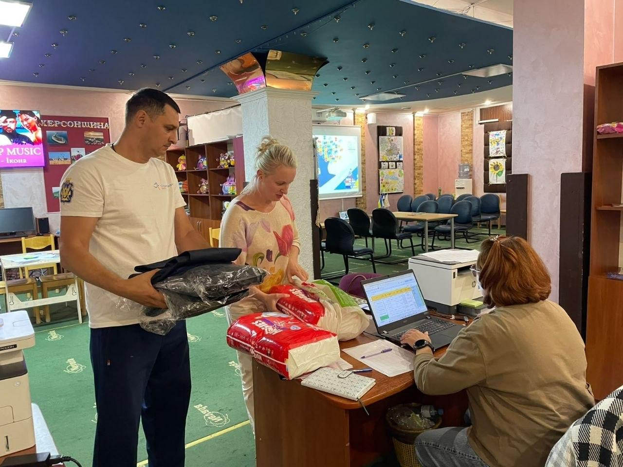 Чоловік та жінка отримують гуманітарну допомогу в приміщенні хабу, де організовано пункт видачі. Ліворуч стоїть чоловік у білій футболці та синіх спортивних штанах, який тримає в руках упакований у плівку теплий одяг або ковдру. Поруч із ним жінка у світлій кофті з квітковим принтом розкладає на робочому столі великі пакунки з підгузками Huggies червоного кольору. Праворуч за цим столом сидить співробітниця хабу в коричневій кофті, яка вносить дані відвідувачів у ноутбук та звіряє інформацію з електронною таблицею. На задньому плані видно залу з синіми стільцями, патріотичними плакатами «Херсонщина» та великим інформаційним екраном. Видача речей організована в межах всебічної підтримки переселенців із Херсонщини в координаційному центрі «Вільні разом» в Ужгороді, де відвідувачі простору отримали 2 набори засобів гігієни, що спрямовано на забезпечення базових побутових потреб і підтримку сімей із дітьми ВПО.
