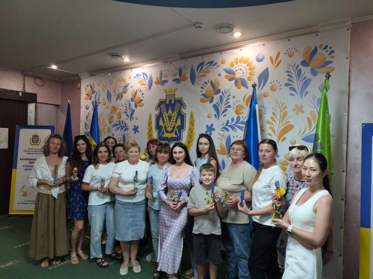 Велике спільне фото групи жінок та хлопчика, які позують в інтер'єрі хабу на тлі стіни, яскраво розписаної традиційним українським квітковим орнаментом у синьо-жовтих тонах із гербом міста Ужгород по центру. Учасники заходу радісно посміхаються та тримають у руках виготовлені власноруч патріотичні синьо-жовті вироби з вощини, прикрашені стрічками. По обидва боки від групи розставлені державні прапори України та прапори міст, а ліворуч видно ролап Херсонської ОВА. Творчий захід організовано в межах всебічної підтримки переселенців із Херсонщини в координаційному центрі «Вільні разом» в Ужгороді, де 12 ВПО взяли участь в арттерапевтичному занятті «Патріотична свічка з вощини своїми руками» з метою надання психологічної допомоги, емоційного розвантаження, згуртування громади та збереження народних традицій.