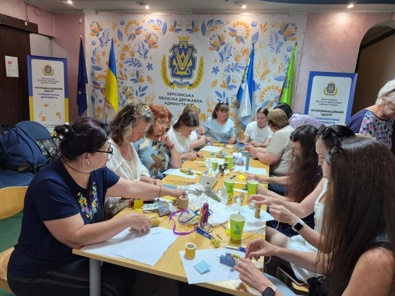 Група жінок різного віку сидить навколо довгого спільного столу в залі хабу та зосереджено виготовляє вироби ручної роботи під час психологічного тренінгу. Учасниці зустрічі працюють із вощиною, нитками, кольоровим папером та сухоцвітами, розкладаючи творчі матеріали, ножиці та паперові стаканчики на столі. На задньому плані залу висить великий банер із офіційним гербом Херсонської обласної державної адміністрації на тлі яскравого синьо-жовтого петриківського розпису, а по обидва боки від нього встановлено державні прапори України та прапори громад. Праворуч і ліворуч також розміщено інформаційні ролапи координаційного центру. Психосоціальний захід організовано в межах всебічної підтримки переселенців із Херсонської області в координаційному центрі «Вільні разом» в Ужгороді, де для 12 переселенок провели зустріч «Коло підтримки: простір для відновлення та внутрішнього ресурсу» з метою надання психологічної допомоги, емоційного розвантаження, пошуку внутрішніх сил, соціальної адаптації та підтримки ментального здоров'я жінок ВПО.