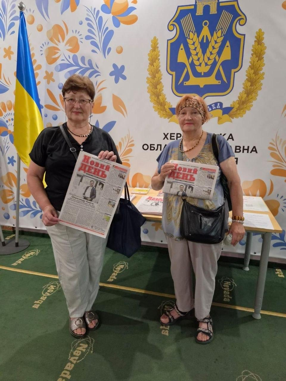 Дві жінки старшого віку позують в інтер'єрі хабу на тлі великого банера з гербом Херсонської ОДА та петриківським розписом. Жінки тримають у руках примірники херсонської газети «Новий день», а ліворуч від них встановлено державний прапор України. Роздача друкованої преси організована в межах всебічної підтримки переселенців із Херсонської області в координаційному центрі «Вільні разом» в Ужгороді, де серед відвідувачів простору поширили 53 примірники обласної газети Херсонщини «Новий день» задля інформування про новини рідного краю, подолання інформаційного вакууму та підтримки зв'язку з домом.
