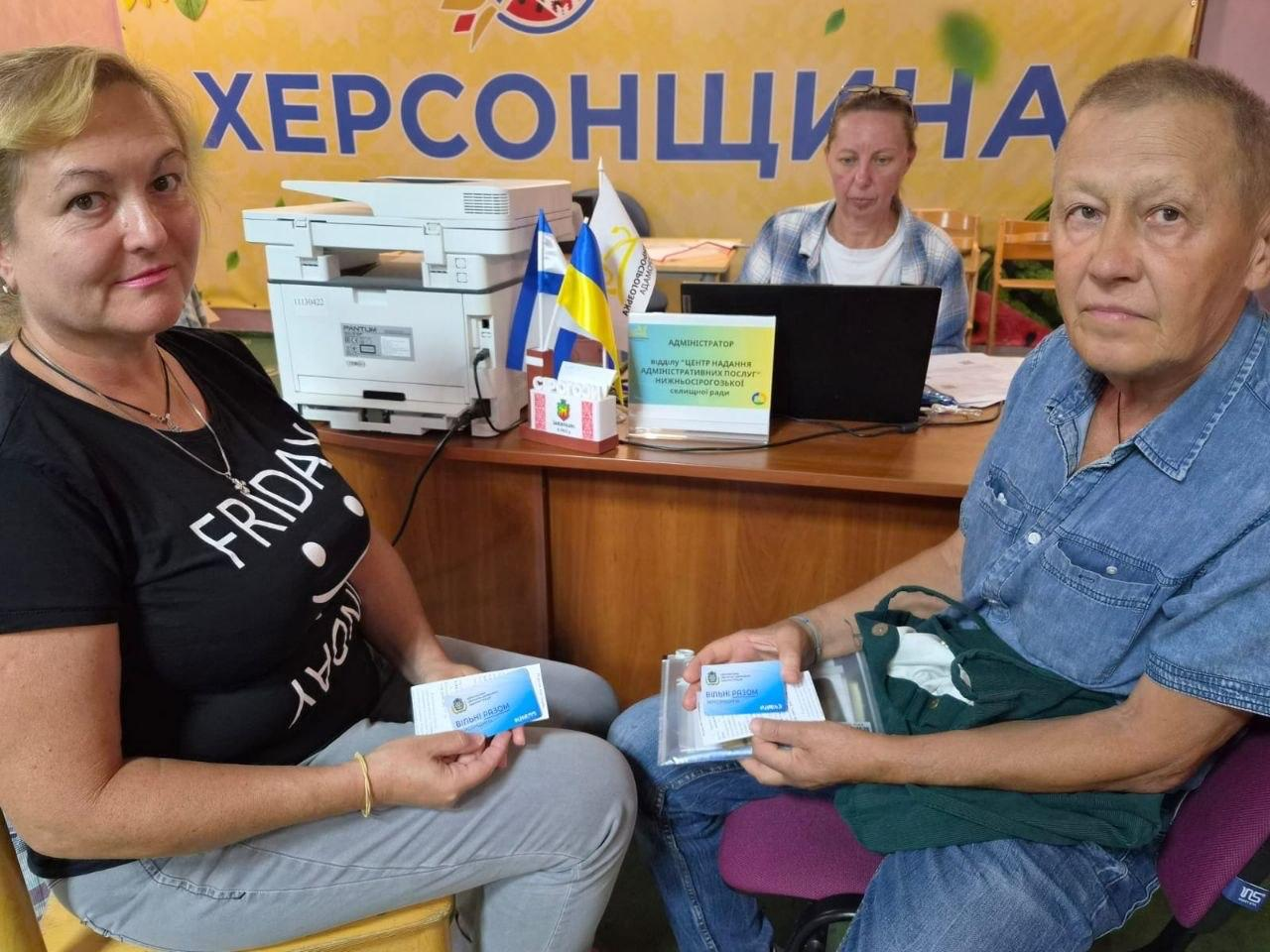 Чоловік та жінка сидять перед робочим столом у хабі та тримають у руках отримані пластикові картки блакитного кольору із символікою координаційного центру. Жінка ліворуч одягнена в чорну футболку з написом, а чоловік праворуч — у синю джинсову сорочку. На задньому плані за столом із ноутбуком та БФП сидить співробітниця хабу, а поруч розміщено табличку адміністратора відділу ЦНАП Нижньосірогозької селищної ради та настільні прапорці. Над робочим місцем висить великий банер із написом «Херсонщина». Надання допомоги організоване в межах всебічної підтримки переселенців із Херсонської області в координаційному центрі «Вільні разом» в Ужгороді, де відділ ЦНАП надав 9 адмінпослуг та 44 консультації з питань адміністративних та публічних послуг задля швидкого розв'язання насущних юридичних та побутових питань внутрішньо переміщених осіб.