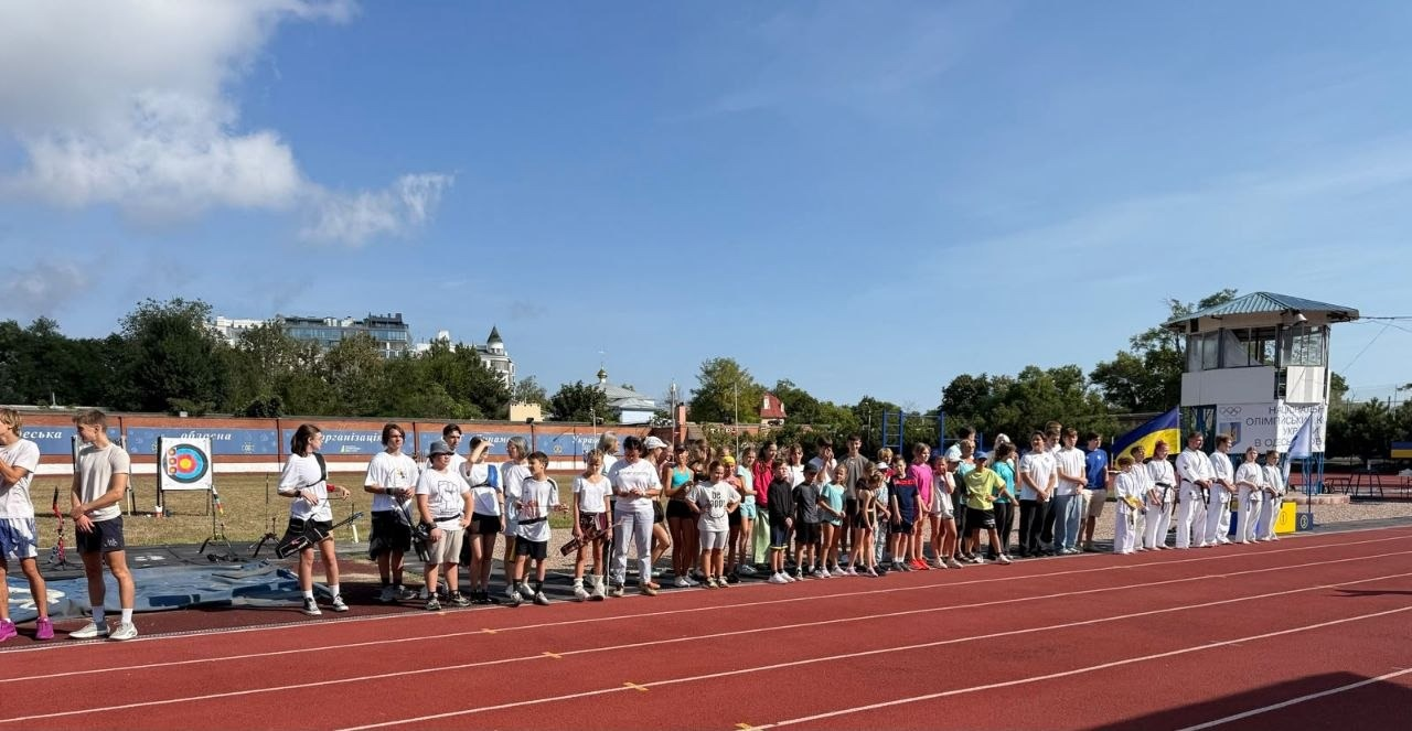 Діти-переселенці з Херсонщини та вихованці одеської організації ФСТ «Динамо» України вишикувалися на червоній біговій доріжці відкритого стадіону в Одесі під час спортивного свята. Учасники, чий приїзд організував центр «Вільні разом», одягнені у світлу спортивну форму, а праворуч стоять юнаки у білих кімоно. Зліва на передньому плані зафіксовані діти зі спортивними луками на тлі встановленої на полі круглої мішені. На задньому плані в сонячний день на тлі дерев видно білу суддівську вишку, державний прапор України та білий банер.
