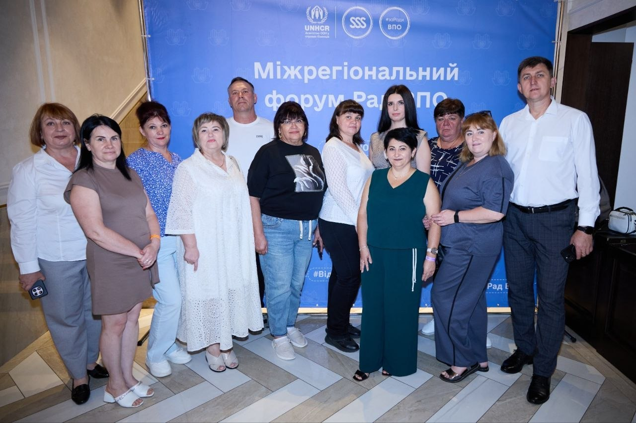 Група представників Рад ВПО Херсонської області позує для спільного фото в Одесі на тлі великого синього інформаційного банера під час Південноукраїнського міжрегіонального форуму Рад ВПО. Дванадцять учасників заходу стоять разом, посміхаючись у кадр. На задньому плані встановлено брендвол із великим білим написом «Міжрегіональний форум Рад ВПО», у верхній частині якого нанесено офіційні логотипи, зокрема Агентства ООН у справах біженців УВКБ ООН. Делегати одягнені у повсякденний літній одяг, а по центру на передньому плані виділяється жінка в довгому темно-зеленому комбінезоні. Присутні зафіксовані на світлій кахельній підлозі, що викладена візерунком у ялинку.