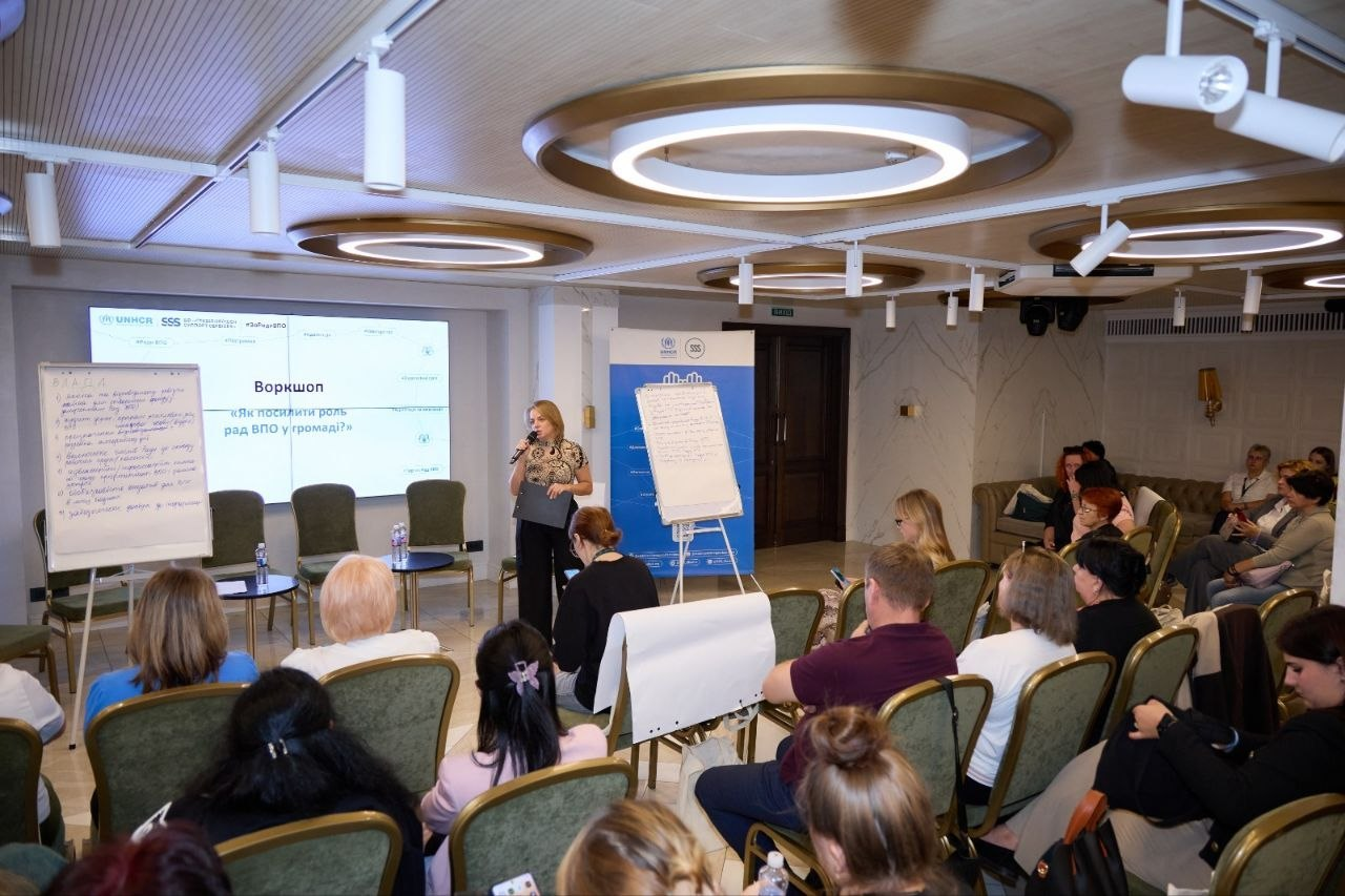Жінка-спікерка виступає з мікрофоном у руках перед аудиторією в Одесі під час Південноукраїнського міжрегіонального форуму Рад ВПО. Вона одягнена у темні штани та блузу з візерунком, тримає папку з документами й стоїть по центру зали біля великого настінного екрана, де транслюється заставка з написом «Воркшоп «Як посилити роль рад ВПО у громаді?»». Учасники заходу, серед яких делегація Херсонської області та очільниці Рад ВПО Анна Мінько, Юлія Чайковська, Наталія Шрам і Маргарита Гришко, сидять півколом на зелених стільцях та слухають доповідь. У залі встановлено два білі фліпчарти з робочими нотатками та синій інформаційний банер у глибині. Сучасне приміщення обладнане оригінальними круглими світильниками на стелі та трековими прожекторами.