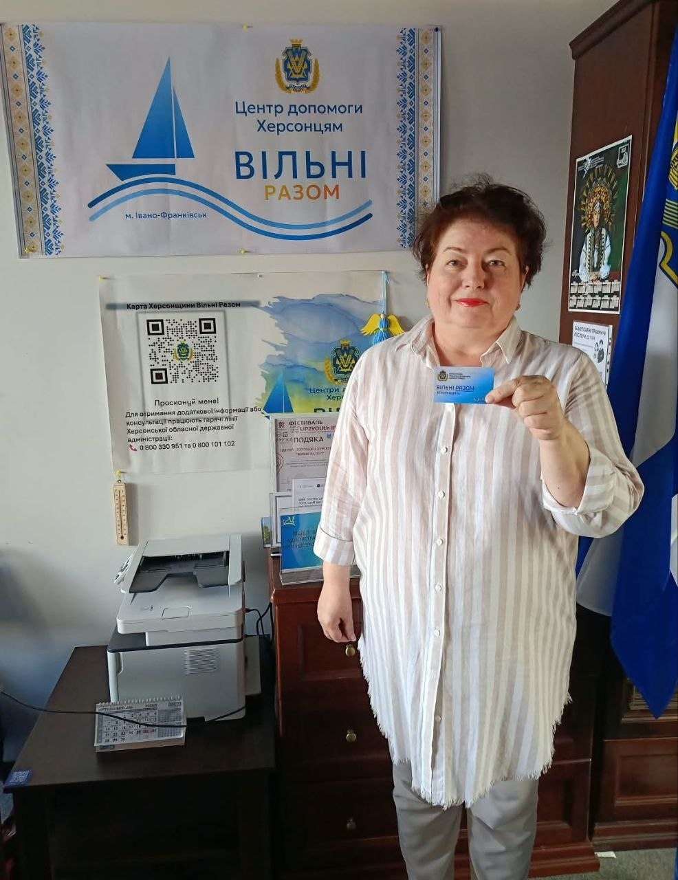 Жінка середнього віку з короткою темною зачіскою позує для фото в івано-франківському координаційному центрі «Вільні разом» під час реалізації проєкту «Картка Херсонщини». Вона одягнена у світлу подовжену сорочку в тонку вертикальну смужку та сірі штани, посміхається в кадр і тримає у лівій руці отриману пластикову картку з логотипом центру. На задньому плані над нею висить великий білий офіційний банер із синім силуетом корабля та написом «Центр допомоги Херсонцям ВІЛЬНІ РАЗОМ м. Івано-Франківськ». Ліворуч на стіні під банером розміщено інформаційний плакат із великим QR-кодом і номерами гарячої лінії Херсонської ОДА. Також у кабінеті видно офісний стіл із принтером, державний прапор України та дерев'яну шафу.