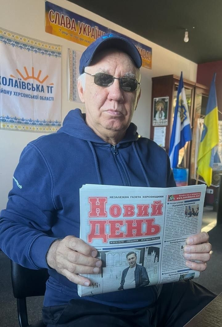 Чоловік похилого віку, переселенець із Херсонщини, тримає в руках свіжий випуск обласної газети в івано-франківському координаційному центрі «Вільні разом». Він одягнений у синю спортивну кофту-худі, темну кепку та сонцезахисні окуляри. Чоловік сидить за столом і розгортає перед собою незалежну газету Херсонщини «Новий день», на першій шпальті якої надруковано великий червоний заголовок та фотографію з офіційного заходу. На задньому плані ліворуч на стіні висить інформаційний плакат із символікою та написом «Миколаївська ТГ району Херсонської області», а вище — патріотичне гасло «Слава Україні!». У правому кутку кімнати на тлі дерев'яних меблів встановлено державний прапор України та офіційний синьо-жовтий стяг Херсонської області.