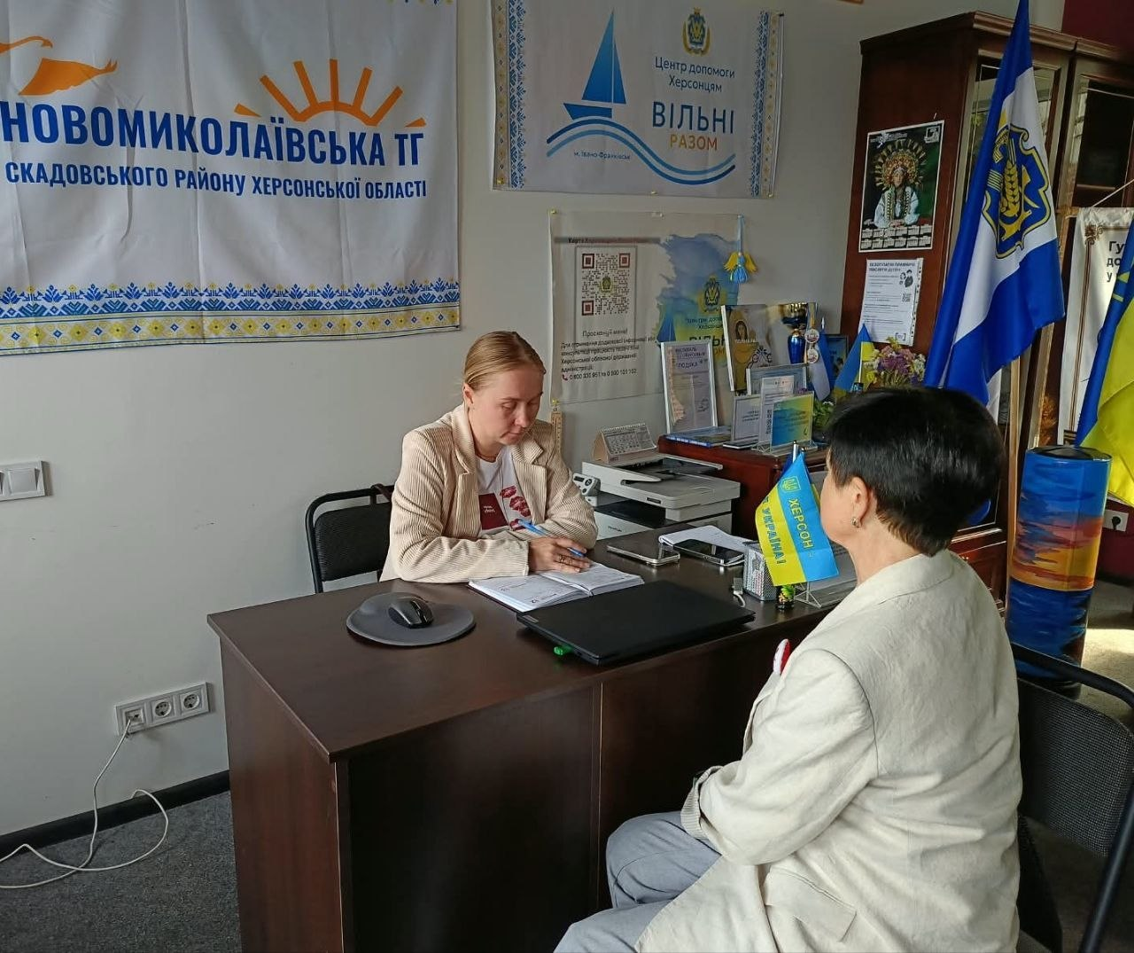Фахівець ЦНАПу надає адміністративні послуги відвідувачці за робочим столом в офісному кабінеті івано-франківського координаційного центру «Вільні разом». Жінка-консультантка у світлому жакеті сидить ліворуч і робить записи у журналі, приймаючи жителів Новомиколаївської, Нижньосірогозької та Каховської громад. Навпроти неї сидітиме літня жінка, яка звернулася за підтримкою. На столі встановлено ноутбук та прапорці України і Херсона. На стіні ліворуч висить банер «Новомиколаївська ТГ Скадовського району Херсонської області», а поруч розміщено логотип центру «Вільні разом» та мапу України. У кутку кабінету стоять великі державний та обласний прапори.
