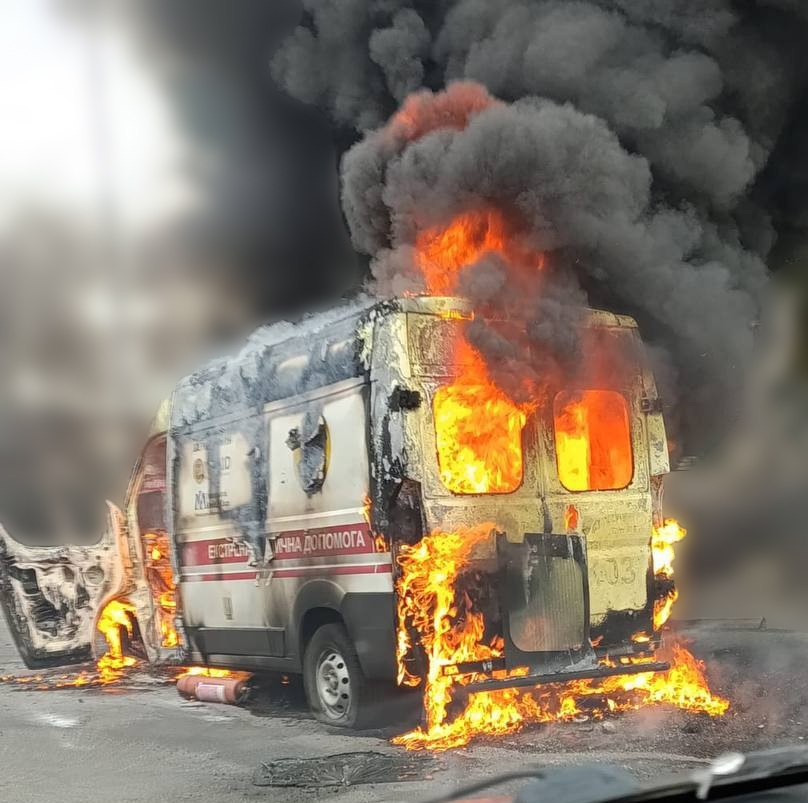 Автомобіль швидкої медичної допомоги, охоплений потужним вогнем на дорозі у Корабельному районі Херсона після прямого влучання російського безпілотника. Спецавтомобіль білого кольору з червоною горизонтальною смугою та написом «ЕКСТРЕНА МЕДИЧНА ДОПОМОГА» зафіксований у момент пожежі з відкритими передніми дверима. Яскраве помаранчеве полум'я повністю охопило задню та внутрішню частини салону, вириваючись із розбитих вікон дверей, а над дахом машини підіймається величезний стовп густого чорного диму. На асфальті поруч із пошкодженим колесом лежить використаний вогнегасник.