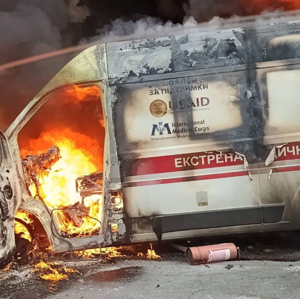 Автомобіль швидкої медичної допомоги, зафіксований крупним планом у момент пожежі після прямого влучання російського безпілотника у Корабельному районі Херсона. Кабіна водія повністю охоплена яскравим помаранчевим полум'ям, яке знищує внутрішнє обладнання, а внаслідок терористичної атаки транспортний засіб вигорів ущент. На вцілілій бічній частині білого кузова з червоною смугою чітко видно напис «ЕКСТРЕНА МЕДИЧНА ДОПОМОГА», а також логотипи міжнародних партнерів: напис «ОНОВЛЕНО ЗА ПІДТРИМКИ USAID» та емблему «International Medical Corps» (Міжнародний Медичний Корпус). Кузов машини вкритий кіптявою та обгорілою фарбою, а на асфальті перед відкритими дверима лежить використаний вогнегасник.