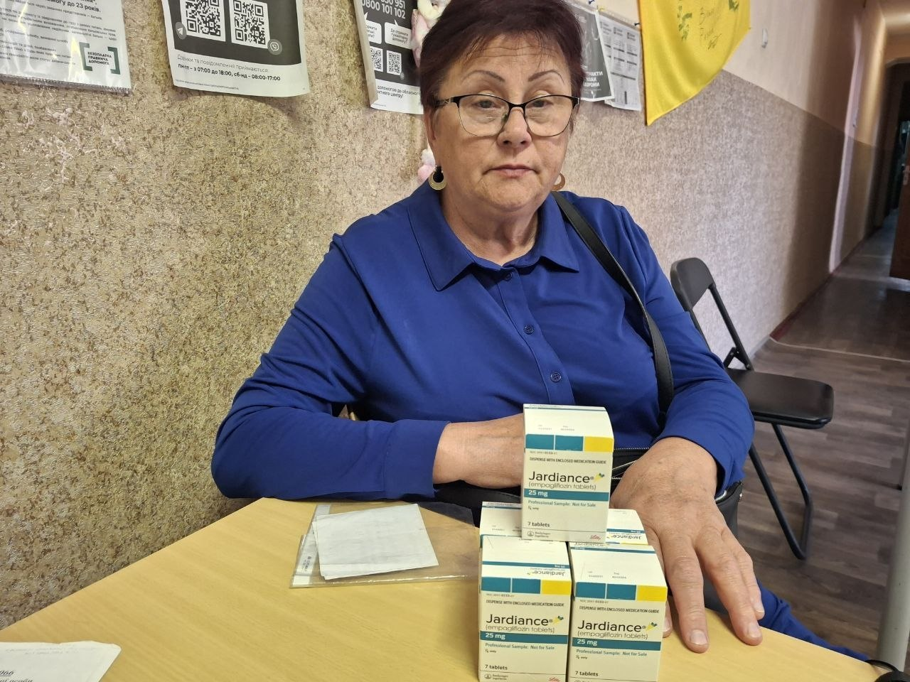 Жінка середнього віку в окулярах та синій сорочці сидить за дерев'яним столом у хмельницькому координаційному центрі «Вільні разом» під час видачі безкоштовних медикаментів внутрішньо переміщеним особам із Херсонщини. Перед нею на столі розкладено чотири біло-жовті упаковки лікарського засобу, які є частиною гуманітарної допомоги з-поміж 67 одиниць лікарських засобів. На задньому плані вздовж світло-бежевої стіни коридору розміщено інформаційні плакати з QR-кодами, оголошення та офіційний розклад роботи хабу.