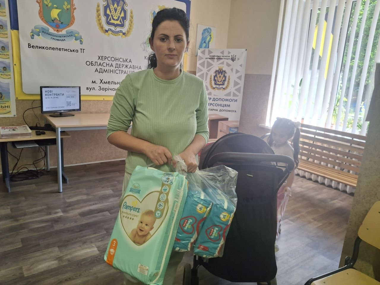 Молода жінка стоїть по центру хмельницького координаційного центру «Вільні разом» під час видачі дитячої гігієни для внутрішньо переміщених осіб із Херсонщини. Вона одягнена в світло-зелений в'язаний джемпер і тримає великі упаковки дитячих підгузків, які є частиною гуманітарної допомоги з-поміж 7 упаковок підгузків. Поруч із жінкою стоїть чорний дитячий візочок із дитиною. На задньому плані ліворуч облаштовано робоче місце з комп'ютером, над яким висять офіційні банери з гербами та написами «Великолепетиська ТГ» і «Херсонська обласна державна адміністрація, м. Хмельницький». Праворуч біля вікна зафіксовано мобільний стенд «Центр допомоги Херсонцям. Гуманітарна допомога».