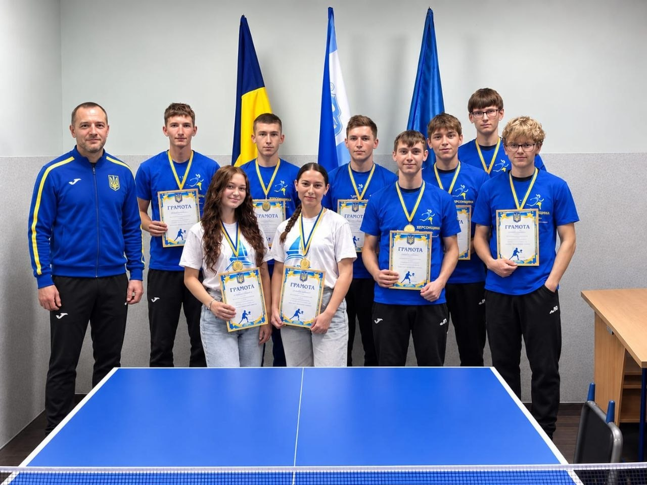 Група юних спортсменів-переселенців із Херсонщини та їхній тренер позують для спільного фото біля синього столу для настільного тенісу після завершення фізкультурно-оздоровчих та спортивно-масових активностей, організованих хмельницьким координаційним центром «Вільні разом». Ліворуч стоїть чоловік-наставник у синій спортивній кофті бренду Joma з гербом України, а поруч із ним вишикувалися семеро хлопців у яскраво-синіх футболках із символікою Херсонщини та медалями на шиях та тримають нагородні грамоти. На передньому плані перед тенісним столом стоять дві дівчини у білих футболках та світлих джинсах, які також тримають отримані нагородні грамоти. На задньому плані за спинами молодіжної команди встановлено три великі прапори: державний стяг України, стяг Херсонської області та частково видно прапор Євросоюзу.