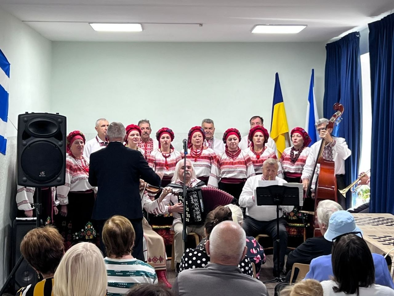Український народний хор та музиканти виступають на сцені у хмельницькому координаційному центрі «Вільні разом» під час урочистого заходу, присвяченого Дню міста Херсона та Дню української музики. Учасники колективу — чоловіки у вишиванках та жінки у традиційних вишитих сорочках і яскраво-червоних головних уборах — стоять півколом на сцені перед глядачами. Перед хористами спиною до кадру стоїть диригент у чорному піджаку, а поруч сидять акомпаніатори, які грають на акордеоні, контрабасі та духових інструментах. На задньому плані праворуч встановлено державний прапор України та стяг Херсонської області, а на стіні ліворуч видно фрагмент прапора міста Херсона. На передньому плані зафіксовані глядачі — внутрішньо переміщені особи з Херсонщини, які слухають святковий концерт.
