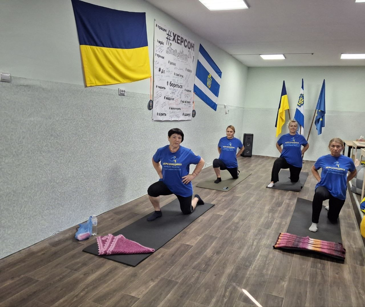 Чотири жінки середнього та старшого віку займаються фітнесом у світлій спортивній залі, виконуючи вправи на індивідуальних килимках-карематах. Учасниці тренування одягнені в однакові сині патріотичні футболки з жовтими написами «Херсонщина. Вільні разом. Спортивний регіон», чорні лосини або спортивні штани та шкарпетки. Вони зафіксовані в момент виконання глибокого випаду на одну ногу, тримаючи руки на поясі для утримання рівноваги. Приміщення має світлі текстурні стіни та дерев'яне покриття підлоги. На стіні ліворуч висить державний прапор України, поруч встановлено великий білий банер із патріотичними хештегами та малюнками міських пам'яток під назвою «#ХЕРСОН», а далі висить прапор міста Херсона. На задньому плані праворуч у кутку зали на флагштоках встановлено три великі стяги: державний прапор України, прапор Херсонської області та прапор Євросоюзу.
