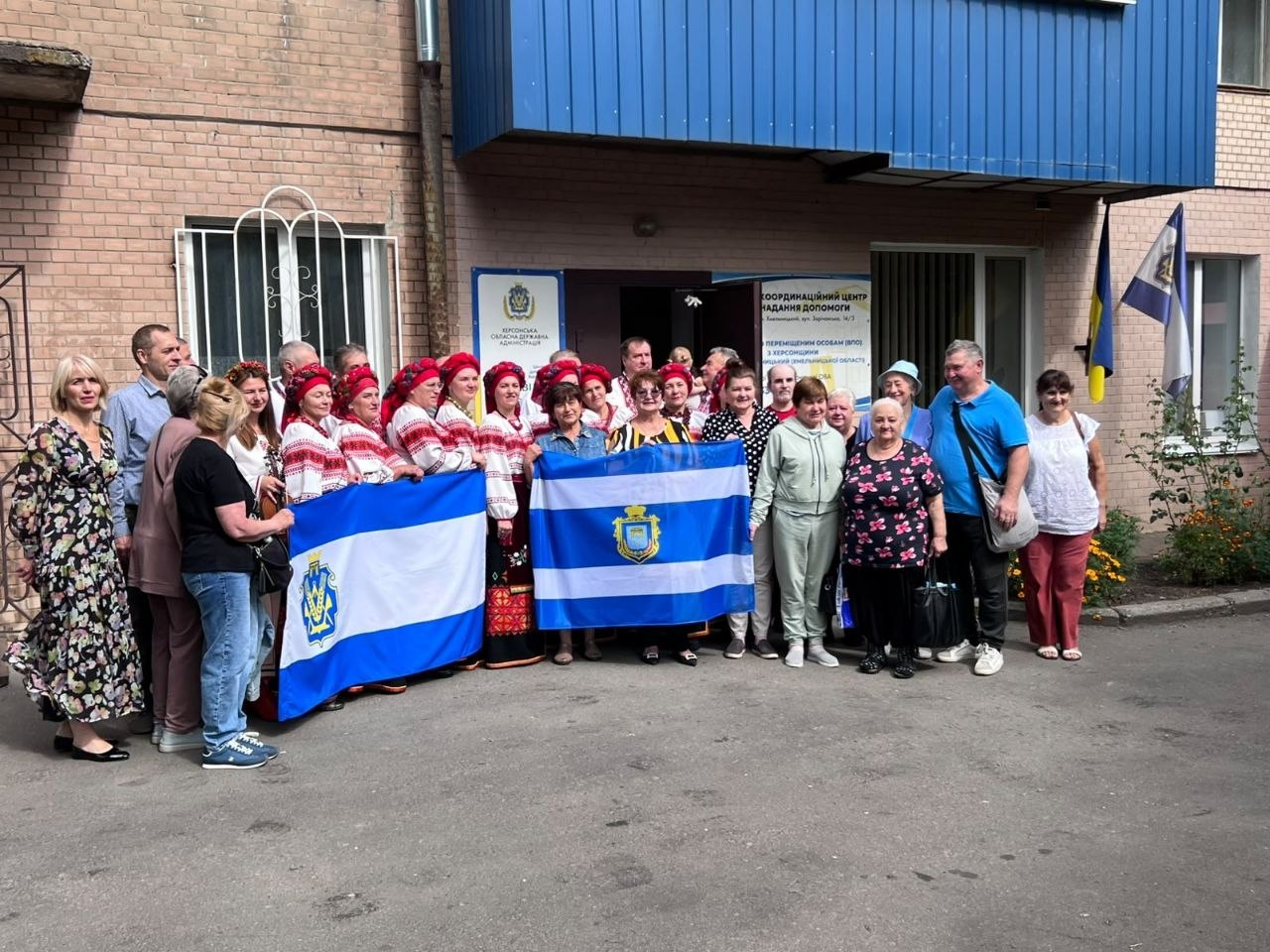 Велика група людей, серед яких учасники народного хору та внутрішньо переміщені особи з Херсонщини, позують для спільного фото на вулиці біля входу до хмельницького координаційного центру «Вільні разом». Присутні чоловіки та жінки різного віку вишикувалися разом на асфальтованому майданчику біля цегляної будівлі хабу, тримаючи перед собою лівіше прапор Херсонської області та правіше прапор міста Херсона. З лівого боку кадру стоять жінки з хору, одягнені в традиційні українські вишиванки, довгі спідниці та червоні головні убори з намистом. Інші переселенці одягнені в повсякденний літній одяг: сукні, футболки, сорочки та штани. На задньому плані біля дверей центру встановлено інформаційні банери: ліворуч — «Херсонська обласна державна адміністрація», а праворуч — «Координаційний центр надання допомоги переміщеним особам (ВПО) з Херсонщини». Біля правого краю будівлі на флагштоках встановлено державний прапор України та прапор Херсонської області.