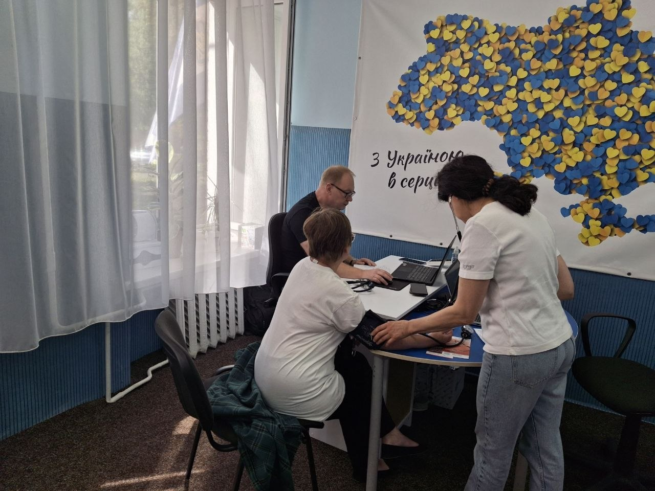 Медпрацівниця вимірює артеріальний тиск жінці у хмельницькому координаційному центрі «Вільні разом» під час прийому мобільної бригади Товариства Червоного Хреста України. На передньому плані праворуч спиною до кадру стоїть медсестра у білій футболці та блакитних джинсах, яка закріплює манжету тонометра на плечі відвідувачки з-поміж ВПО з Херсонщини. Пацієнтка сидить на стільці боком до кадру у білій футболці, а за цим же столом по центру сидить чоловік-лікар в окулярах та чорній футболці, який вносить дані пацієнтки у ноутбук, надаючи медичну консультацію. Кабінет має сині панелі на стінах та велике вікно ліворуч із білою прозорою тюлю. На стіні праворуч встановлено великий білий банер із написом «З Україною в серці», на якому зображено карту України, викладену з маленьких жовтих та блакитних патріотичних сердечок.