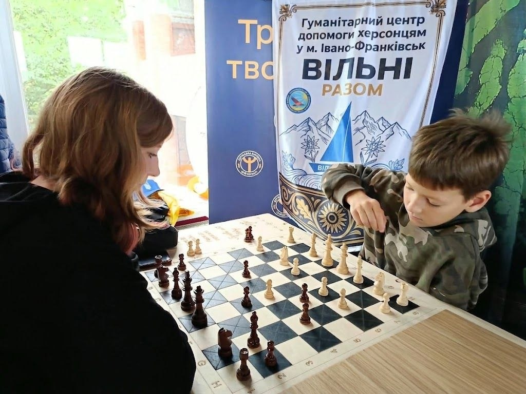 Хлопчик та дівчина грають у шахи за столом в івано-франківському гуманітарному центрі «Вільні разом» під час чемпіонату з настільних ігор, організованого до Тижня фізичної культури. [Freshness] Справа на передньому плані зафіксований маленький хлопчик у камуфляжній кофті, який зосереджено обмірковує хід, тримаючи руку біля білої шахової фігури на дошці. Навпроти нього ліворуч спиною до кадру сидітиме дівчина з русявим волоссям, яка грає темними фігурами. Змагання зібрало внутрішньо переміщених осіб із Херсонської та Харківської областей, а також місцевих жителів і військовослужбовців. На задньому плані за столом встановлено великий офіційний білий банер із синім силуетом корабля та написом «Гуманітарний центр допомоги херсонцям у м. Івано-Франківськ ВІЛЬНІ РАЗОМ».