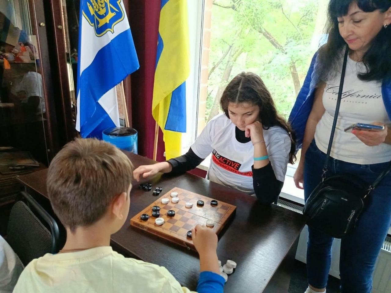 Двоє юних учасників змагаються в шашки за столом в івано-франківському гуманітарному центрі «Вільні разом» під час чемпіонату з настільних ігор. На передньому плані спиною до кадру сидить хлопчик у світло-жовтій футболці, який обмірковує свій хід білими фігурами. Навпроти нього зафіксовано дівчину з довгим темним волоссям у білій патріотичній футболці з написом «#BEACTIVE | EUROPEAN WEEK OF SPORT», яка зосереджено підперла рукою підборіддя та грає чорними шашками. Праворуч за поєдинком уважно спостерігає виконавча директорка Херсонського обласного відділення НОК України Луїза Кірсанова. На задньому плані біля вікна на флагштоках встановлено державний прапор України та стяг Херсонської області.