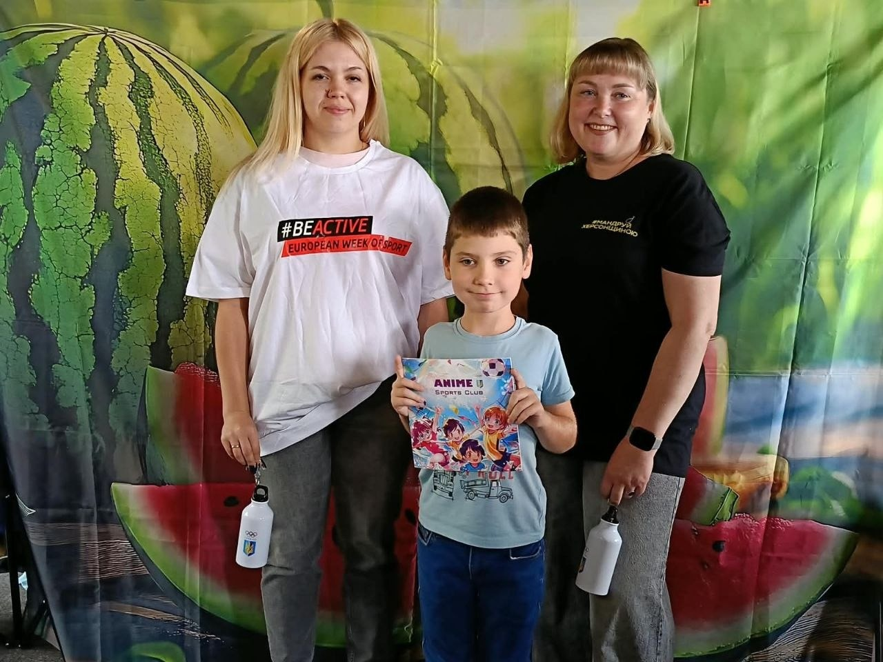 Маленький хлопчик та дві жінки-організаторки позують для фото в івано-франківському координаційному центрі «Вільні разом» на тлі патріотичної фотозони з великим херсонським кавуном. По центру стоїть дитина у блакитній футболці та синіх джинсах, яка тримає в руках отриманий пам’ятний подарунок — кольоровий зошит від Херсонського обласного відділення НОК України та Новомиколаївської СВА. Ліворуч від нього стоїть дівчина у білій футболці з логотипом «#BEACTIVE | EUROPEAN WEEK OF SPORT» та сірих джинсах, а праворуч — жінка у чорній футболці з написом «МАНДРУЙ ХЕРСОНЩИНОЮ». Обидві організаторки тримають у руках подарункові спортивні пляшки для води білого кольору. На задньому плані облаштовано яскравий банер із зображенням скибок стиглого кавуна.