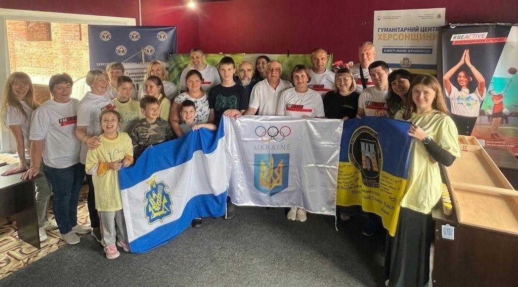 Велика група учасників та організаторів позує для спільного фото в івано-франківському гуманітарному центрі «Вільні разом» під час чемпіонату з настільних ігор, присвяченого Тижню фізичної культури і спорту. Двадцять три людини різного віку — чоловіки, жінки, діти та військовослужбовці НГУ — вишикувалися в кілька рядів, посміхаючись у кадр. По центру присутні тримають три великі стяги: ліворуч — біло-синій прапор Херсонської області, посередині — білий прапор Національного олімпійського комітету України з олімпійськими кільцями та тризубом, а праворуч — синьо-жовтий прапор із символікою підрозділу. Учасники змагань одягнені переважно у білі та світлі патріотичні футболки. На задньому плані вздовж бордової стіни встановлено банер гуманітарного центру Херсонщини та вертикальний ролап «#BEACTIVE | EUROPEAN WEEK OF SPORT», а праворуч видно дерев'яну настільну дошку для джакколо.