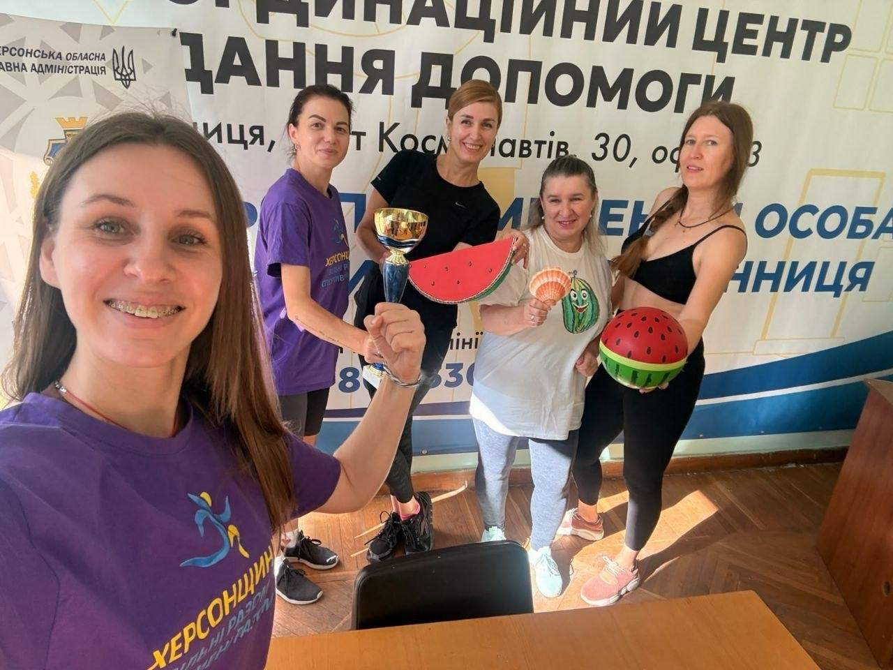 П'ять жінок-переселенок із Херсонщини позують для спільного селфі у вінницькому координаційному центрі «Вільні разом» під час Тижня фізичної культури і спорту. На передньому плані ліворуч зафіксовано жінку у фіолетовій патріотичній футболці «Херсонщина. Вільні разом», яка робить знімок. Позаду неї стоять ще чотири учасниці заходу, які щиро посміхаються після завершення патріотичного тренування. Жінки тримають у руках тематичні сувеніри та інвентар: спортивний кубок, декоративні вироби у вигляді скибок кавуна, розмальовану мушлю та круглий м'яч, стилізований під кавун. На задньому плані встановлено великий біло-синій банер із написами «КООРДИНАЦІЙНИЙ ЦЕНТР НАДАННЯ ДОПОМОГИ ПЕРЕМІЩЕНИМ ОСОБАМ ХЕРСОНЩИНА», де вказано офіційну адресу у Вінниці.