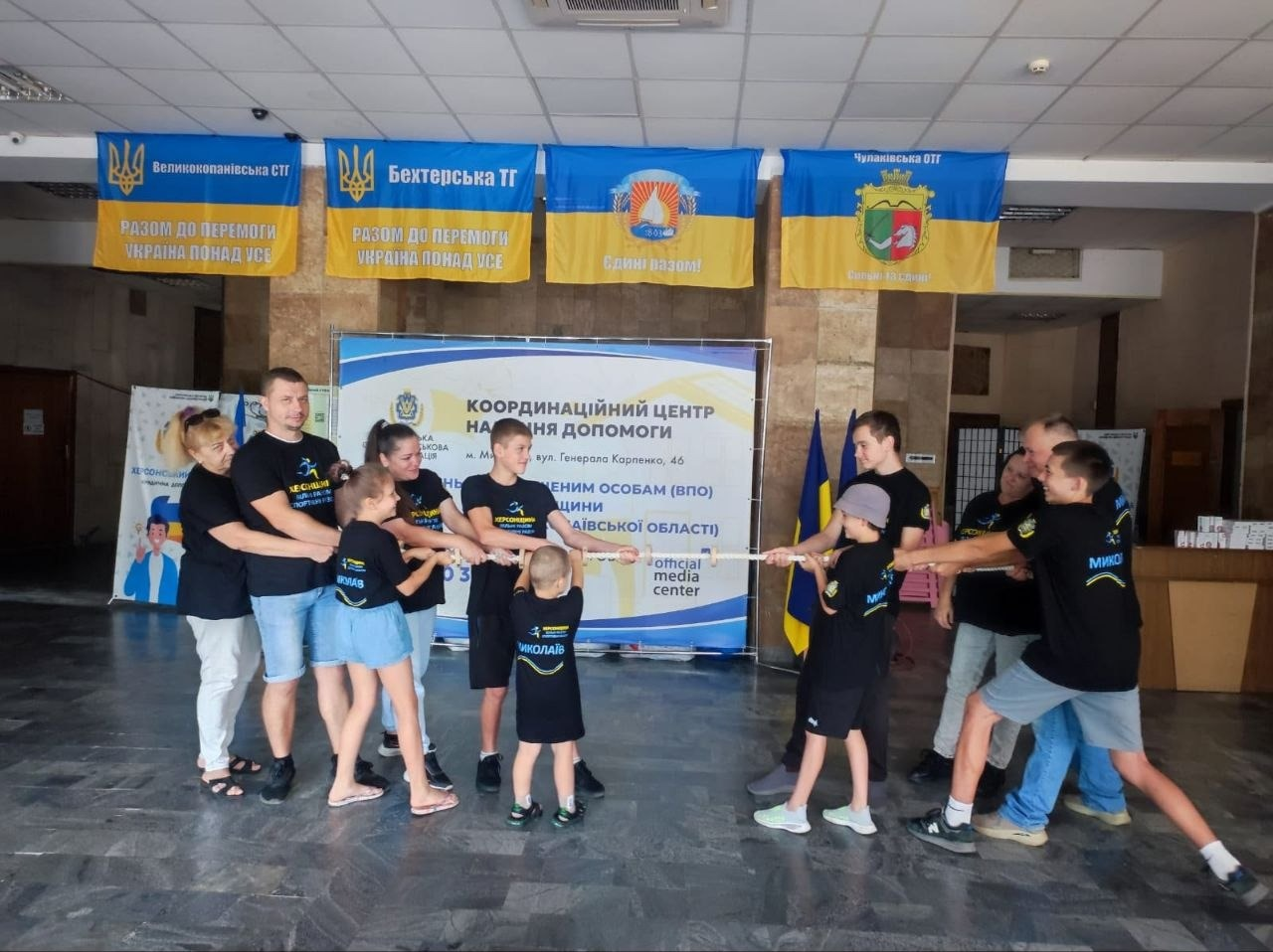 Дві команди дорослих та дітей змагаються з перетягування каната під час сімейних вихідних «Вільні, спортивні, єдині!» в миколаївському координаційному центрі «Вільні разом». Учасники обох команд — переселенці з Лазурненської, Бехтерської, Чулаківської та Великокопанівської громад у чорних патріотичних футболках — міцно тримають товстий білий канат та із зусиллям тягнуть його у свої боки під час «веселих стартів» до Дня фізичної культури і спорту. На задньому плані встановлено великий білий банер координаційного центру допомоги ВПО з Херсонської області у місті Миколаїв (вул. Генерала Карпенка, 46). Над банером на стіні розвішані синьо-жовті офіційні прапори Великокопанівської, Бехтерської та Чулаківської територіальних громад.