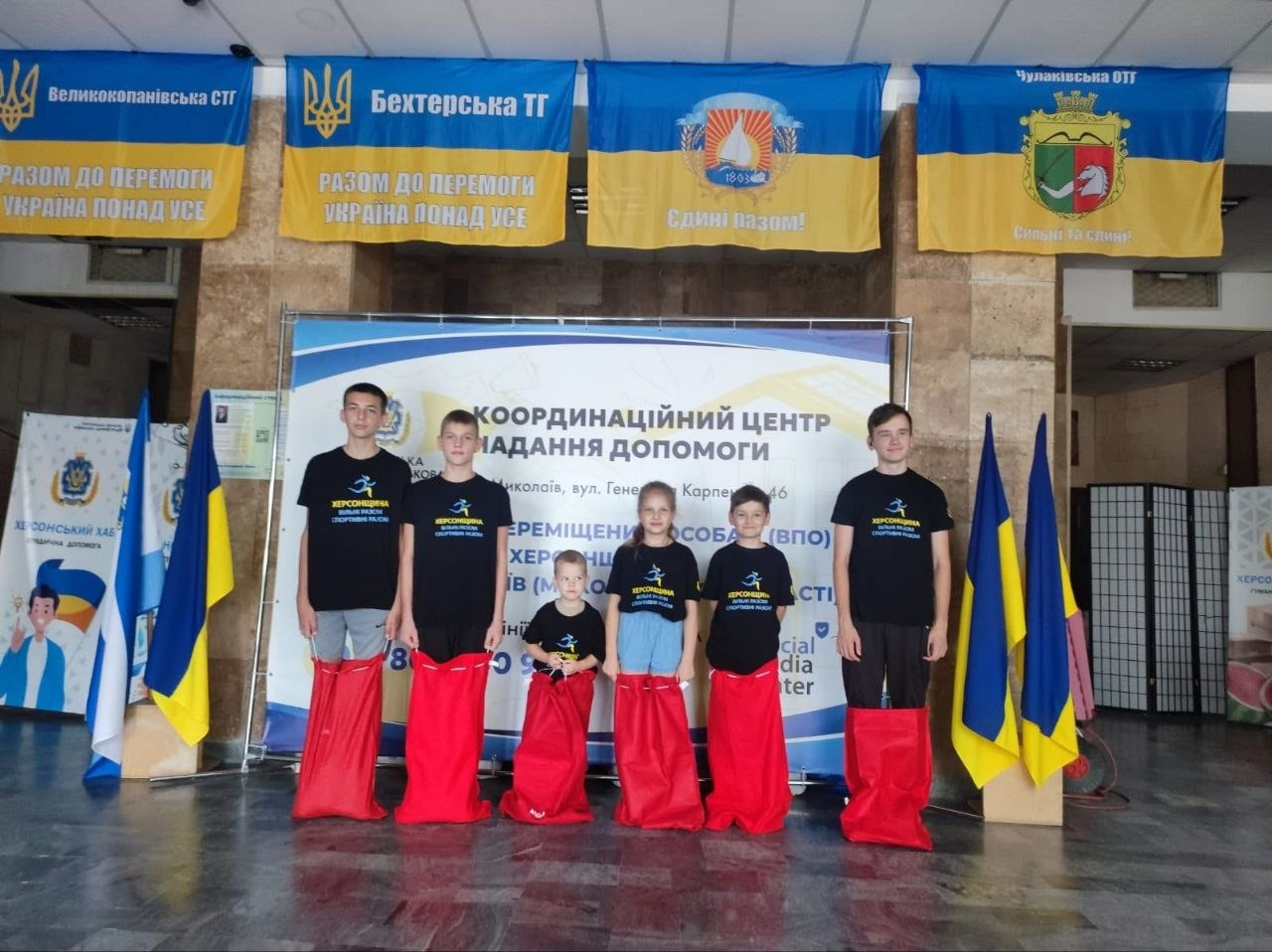 Шість підлітків та дітей позують для фото в холі миколаївського координаційного центру «Вільні разом» під час участі у «веселих стартах» до Дня фізичної культури і спорту. Учасники змагань вишикувалися в один ряд, тримаючи руками на рівні поясу великі яскраво-червоні мішки для стрибків. Діти — переселенці з Лазурненської, Бехтерської, Чулаківської та Великокопанівської громад — одягнені в однакові чорні патріотичні футболки. На передньому плані по центру стоїть найменший хлопчик у футболці з написом «МИКОЛАЇВ». На задньому плані встановлено великий білий банер координаційного центру допомоги ВПО з Херсонщини у місті Миколаїв (вул. Генерала Карпенка, 46). Над банером розвішані чотири офіційні прапори територіальних громад: Великокопанівської, Бехтерської, Чулаківської та Лазурненської, а з обох боків на флагштоках стоять державні прапори України.