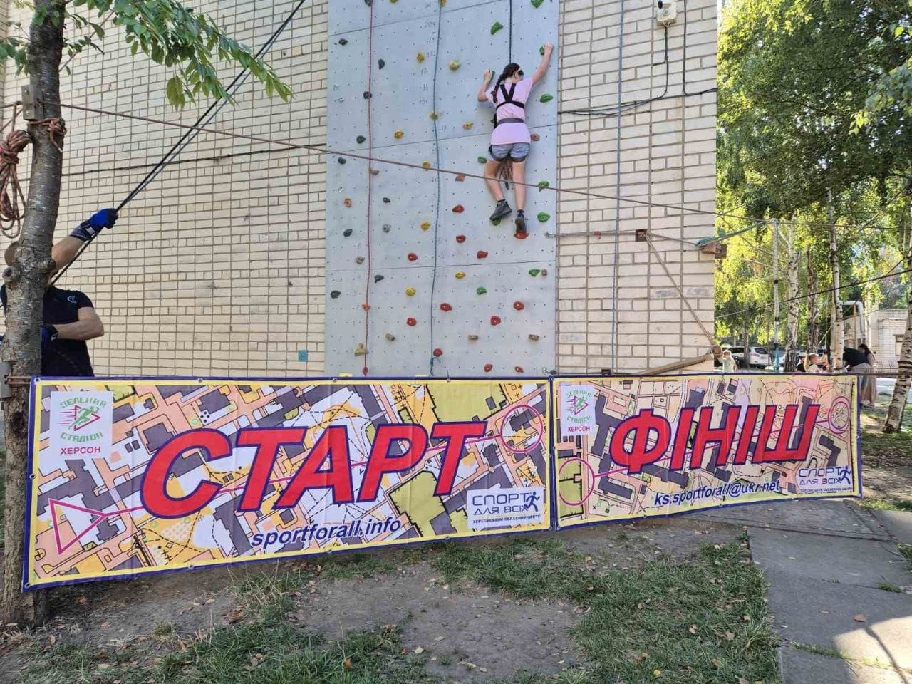 Дівчинка у страхувальному спорядженні підіймається вгору по вуличному скеледрому під час дитячого мінітурніру зі скелелазіння на Херсонщині. Вона зафіксована спиною до кадру на висоті кількох метрів, чіпляючись за спеціальні різнокольорові зачепи на вертикальному щиті, встановленому на цегляній стіні будівлі. Ліворуч біля дерева стоїть тренер-інструктор, який забезпечує страховку за допомогою натягнутого троса під час змагань до Олімпійського дня та Дня фізичної культури і спорту. На передньому плані встановлено великий жовтий банер із великими яскравими написами «СТАРТ» і «ФІНІШ», мапою та офіційними емблемами «Зелений стадіон. Херсон» і «Спорт для всіх». Знімок зроблено в сонячний день на тлі зелених дерев парку.