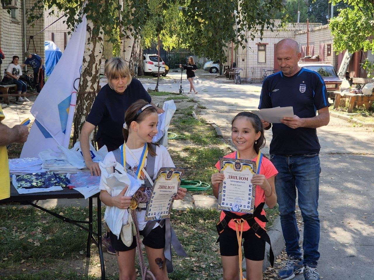 Дві дівчинки-призерки та організатори позують для фото на вулиці під час урочистого нагородження переможців дитячого мінітурніру зі скелелазіння на Херсонщині. Юні спортсменки зі страхувальними системами на поясі щиро посміхаються та тримають у руках отримані офіційні дипломи з медалями на синьо-жовтих стрічках. Ліворуч стоїть дівчинка у білій футболці, а праворуч — її колега у рожевій футболці, які стали переможницями серед 32 учасників із Херсонської громади. За ними праворуч стоїть чоловік-суддя у синій спортивній футболці та джинсах, який переглядає нагородні листи. Ліворуч біля столу з призами та білими пакунками фірмових олімпійських футболок зафіксовано жінку-організаторку. Захід до Олімпійського дня та Дня фізичної культури і спорту проходить на вулиці на тлі цегляної будівлі та зелених дерев.