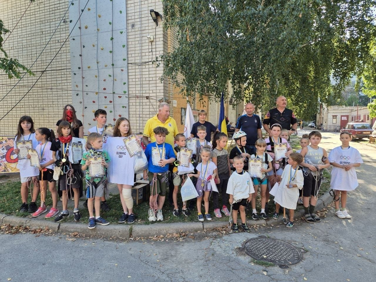 Велика група юних спортсменів та організаторів позує для спільного фото на вулиці після завершення дитячого мінітурніру зі скелелазіння на Херсонщині. Близько двадцяти дітей віком від 6 до 14 років із Херсонської громади вишикувалися в один довгий ряд уздовж бордюру на тлі високої цегляної стіни будівлі, де облаштовано тренувальний скеледром із кольоровими зачепами. Багато учасників тримають у руках отримані офіційні дипломи, медалі та білі фірмові футболки з олімпійською символікою «OLYMPIC DAY». Позаду дітей по центру стоять тренери та організатори змагань, які підтримали проведення турніру до Олімпійського дня та Дня фізичної культури і спорту. Праворуч на задньому плані зафіксовано державний прапор України, зелені дерева та припарковані автомобілі.