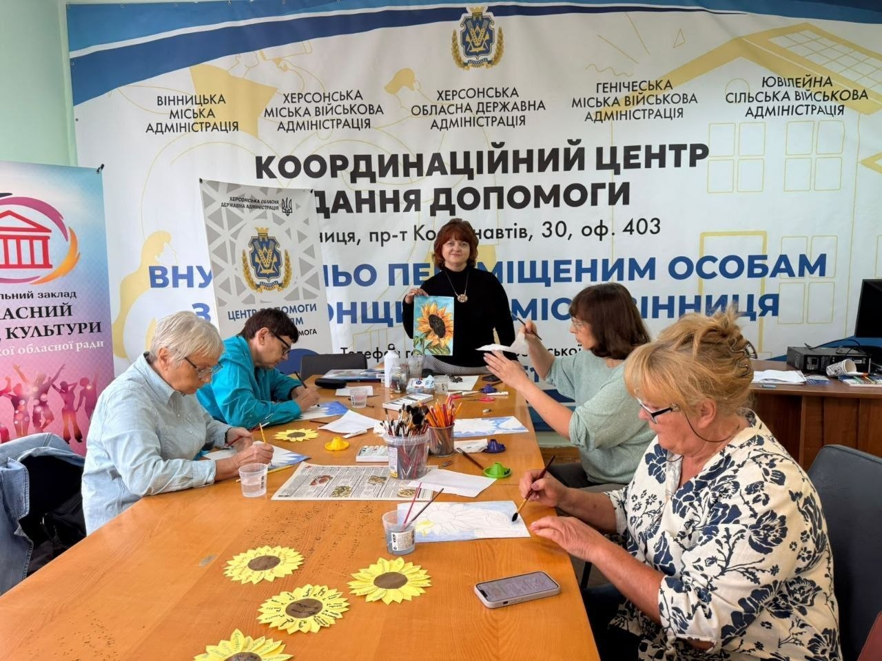 Група літніх жінок-переселенок із Херсонщини займається творчістю у вінницькому координаційному центрі «Вільні разом» під час майстер-класу із малювання в стилі петриківського розпису «Соняхи». П'ять учасниць заходу сидять навколо довгого столу, на якому розкладено фарби, пензлі, склянки з водою та паперові заготовки, захоплено розмальовуючи патріотичні сувеніри. По центру за столом стоїть жінка-модераторка у чорній кофті, яка тримає в руках та демонструє присутнім зразок малюнка із зображенням соняшника. На задньому плані встановлено великий біло-синій банер із написом «КООРДИНАЦІЙНИЙ ЦЕНТР НАДАННЯ ДОПОМОГИ ВНУТРІШНЬО ПЕРЕМІЩЕНИМ ОСОБАМ ХЕРСОНЩИНИ У МІСТІ ВІННИЦЯ» під егідою Херсонської ОВА та міських військових адміністрацій.