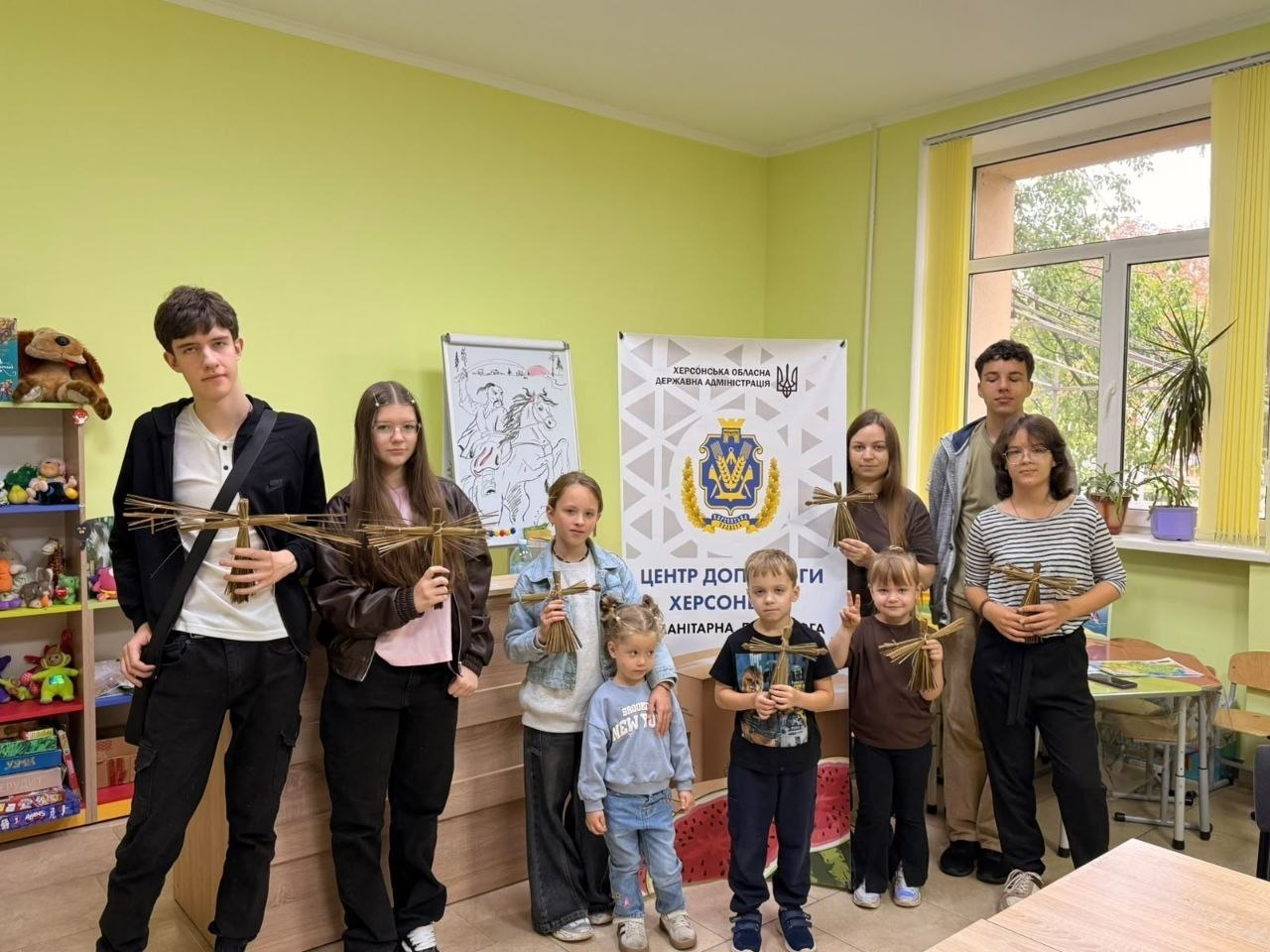 Група дітей, підлітків та молоді позує для спільного фото у вінницькому координаційному центрі «Вільні разом» під час виготовлення сувенірів-оберегів для ЗСУ. Десять учасників заходу різного віку вишикувалися в ряд у світлій кімнаті з ніжно-зеленими стінами, тримаючи в руках власноруч зроблені традиційні українські солом'яні обереги. По центру за дітьми встановлено великий білий банер із гербом та офіційними написами «ХЕРСОНСЬКА ОБЛАСНА ДЕРЖАВНА АДМІНІСТРАЦІЯ» та «ЦЕНТР ДОПОМОГИ ХЕРСОНЦЯМ. ГУМАНІТАРНА ДОПОМОГА». Ліворуч на задньому плані видно дитячий дерев'яний стелаж з іграшками та книгами, а праворуч — велике вікно з кімнатними рослинами.