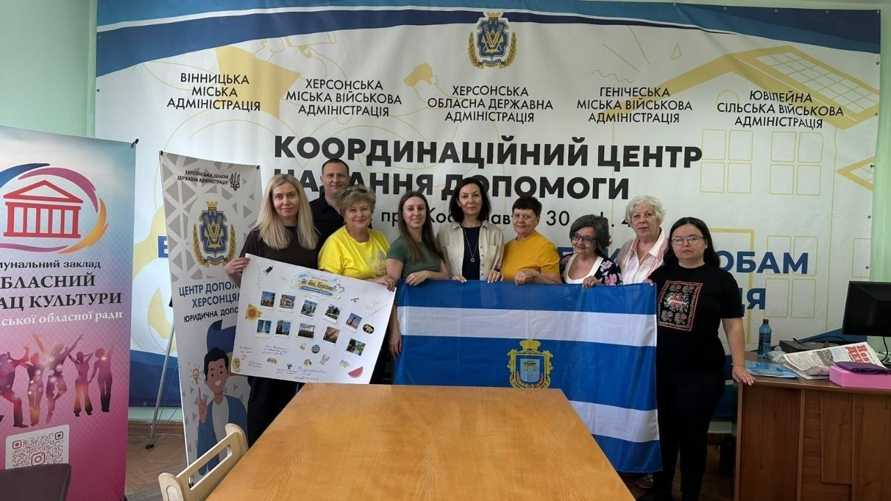 Група переселенців із Херсонщини позує для спільного фото у вінницькому координаційному центрі «Вільні разом» на завершення культурної зустрічі «Як ти, Херсоне?». Десять учасників та організаторів заходу вишикувалися за довгим дерев'яним столом у приміщенні хабу. По центру присутні розгорнули великий біло-синій прапор міста Херсона з гербом, а ліворуч дві жінки тримають спільно оформлений білий фліпбук із фотографіями визначних місць та малюнками херсонських кавунів з написом «Це мій Херсон». Програма, проведена Херсонською обласною бібліотекою для дітей імені Дніпрової Чайки, також передбачала інтерактивний пазл із гербом міста, патріотичну вікторину та частування кавуном. На задньому плані встановлено великий біло-синій банер координаційного центру допомоги ВПО з Херсонщини у місті Вінниця.