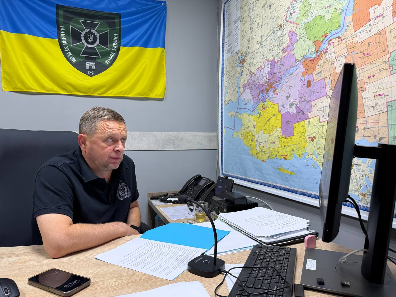 Перший заступник начальника Херсонської ОВА Дмитро Бутрій на робочому місці у своєму кабінеті під час проведення онлайн-зустрічі щодо державної підтримки аграріїв регіону. Посадовець сидить у шкіряному кріслі боком до кадру, одягнений у чорну футболку-поло з шевроном, і зосереджено дивиться на монітор комп'ютера. На дерев'яному столі перед ним розкладено документи у блакитній папці, встановлено настільний мікрофон, лежить смартфон із часом 14:05 на екрані, клавіатура, а також стоять стаціонарні телефони та склянка чаю. На задній сірій стіні ліворуч висить державний прапор України із великим круглим шевроном «Вільний Херсон — Вільна Україна», а праворуч розміщено велику детальну карту Херсонської області з позначенням територіальних громад.