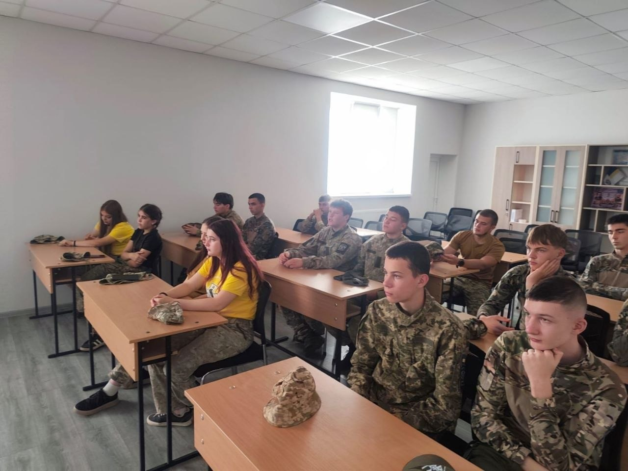 Юні представники Херсонщини з рою «Вільні люди» слухають лекцію в навчальній аудиторії під час всеукраїнського вишколу «Джура-Прикордонник» на Закарпатті. Учні розмістилися по двоє за кількома рядами дерев'яних столів. На передньому плані праворуч зафіксовано двох хлопців у камуфляжних куртках, які зосереджено слухають інструктора. На столах перед деякими підлітками лежать військові кепки-мазепинки. Ліворуч у другому ряду сидить дівчина у патріотичній яскраво-жовтій футболці та камуфляжних штанах. Інші джури в залі також одягнені у військові однострої або футболки. Приміщення кабінету має світлі стіни та велике вікно на задньому плані, а у правому кутку аудиторії встановлено світлу дерев'ну офісну шафу.