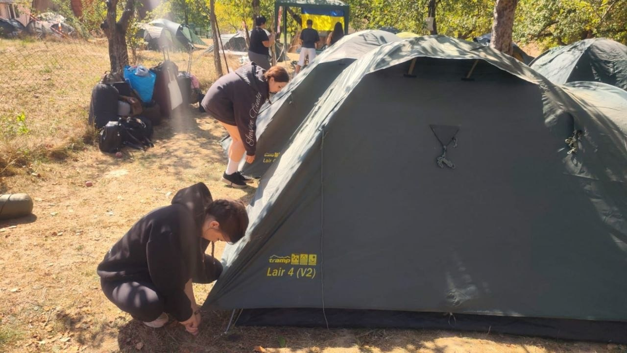 Юні представники Херсонщини з рою «Вільні люди» облаштовують наметове містечко на Закарпатті під час всеукраїнського вишколу «Джура-Прикордонник». На передньому плані ліворуч хлопець у чорному худі присів навпочіпки біля великого чотиримісного туристичного намету темно-зеленого кольору марки «tramp Lair 4 (V2)» та закріплює його основу кілочками до землі. Трохи далі за ним дівчина в темній кофті нахилилася над сусіднім наметом, перевіряючи кріплення. З лівого боку кадру під деревами складено рюкзаки та спальні мішки восьми юних джур, які проходять навчання з орієнтування на місцевості та таборування. На задньому плані сонячної галявини видно інші намети та великий зелений садовий павільйон-альтанку, всередині якого розгорнуто державний прапор України.