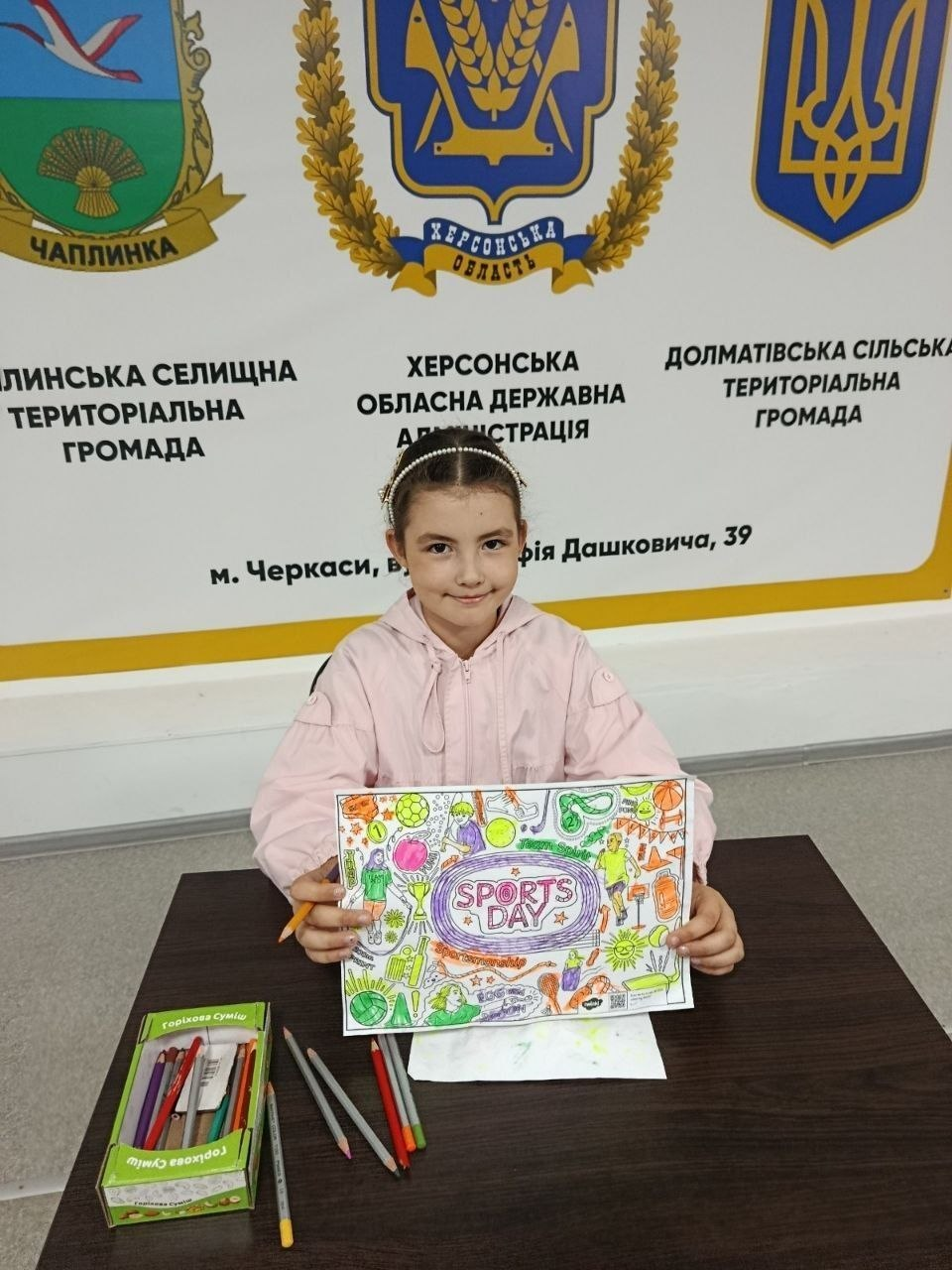 Маленька дівчинка-переселенка з Херсонщини сидить за столом у черкаському координаційному центрі «Вільні разом» та демонструє свій яскравий малюнок під час участі у флешмобі «Мій улюблений вид спорту». Вона одягнена у світло-рожеву кофту та обруч із намистинами, посміхається в кадр і тримає в руках розфарбований олівцями аркуш із великим написом «SPORTS DAY» у центрі, зображеннями м'ячів, спортсменів та олімпійських кілець. Перед нею на темному столі лежить пачка олівців та кілька підточених олівців поруч. Безпосередньо за дівчинкою на стіні встановлено великий білий стенд із гербами та офіційними написами Чаплинської селищної, Долматівської сільської громад та Херсонської ОВА із зазначенням адреси хабу в місті Черкаси.