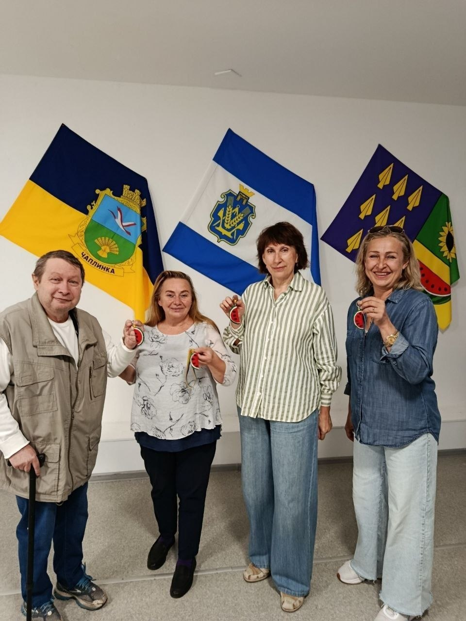 Чотири учасники-переселенці з Херсонщини позують для спільного фото в черкаському координаційному центрі «Вільні разом» на завершення майстерки з виготовлення брелоків-кавунчиків до Дня міста Херсон. Один чоловік та три жінки вишикувалися в ряд на тлі світлої стіни, посміхаючись у кадр і демонструючи виготовлені власноруч сувеніри з повітряного пластиліну. Крайній ліворуч чоловік у світлому жилеті спирається на тростину, а поруч із ним стоять жінки у блузі з квітковим принтом, смугастій та джинсовій сорочках. На задньому плані розвішано три офіційні прапори: синьо-жовтий прапор Чаплинської громади з гербом, біло-синій стяг Херсонської області та прапор Новомиколаївської громади.