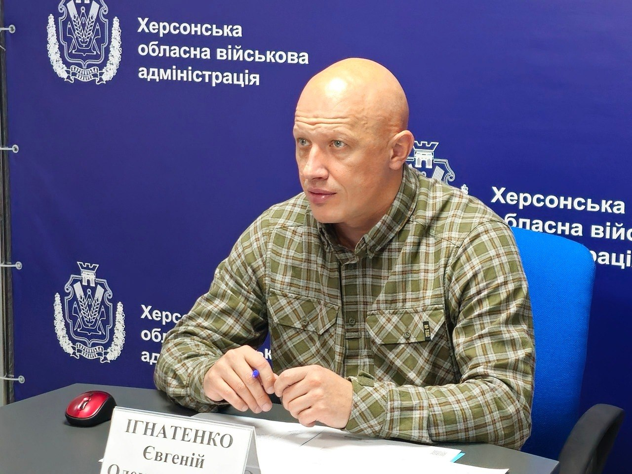 Заступник начальника Херсонської ОВА Євгеній Ігнатенко сидить за робочим столом під час онлайн-наради щодо надання допомоги населенню на опалювальний сезон 2026/2027 років. Посадовець сидить у синьому офісному кріслі на тлі великого яскраво-синього пресволла з повторюваними білими гербами та написами «Херсонська обласна військова адміністрація». Він одягнений у тактичну сорочку в зелено-коричневу клітинку, тримає в руках синю кулькову ручку та зосереджено дивиться перед собою під час обговорення урядової програми підтримки. На сірому столі перед ним лежать документи, розташована комп'ютерна мишка та встановлена  біла настільна табличка з ПІБ посадовця.