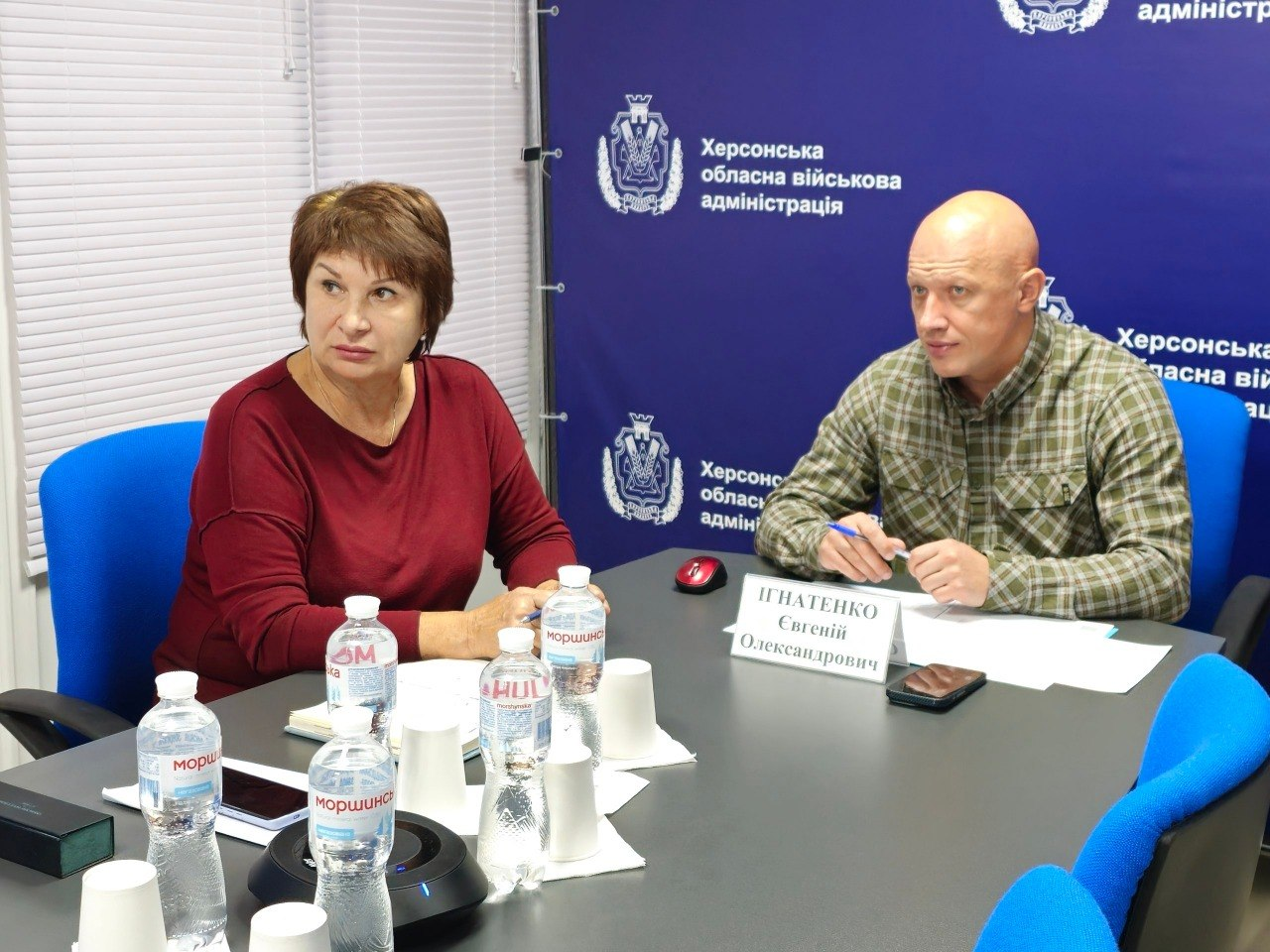 Заступник начальника Херсонської ОВА Євгеній Ігнатенко та заступниця начальника Херсонської МВА Наталія Чорненька сидять за великим столом під час онлайн-наради щодо допомоги населенню на опалювальний сезон 2026/2027 років. Посадовці сидять поруч у синіх офісних кріслах і зосереджено дивляться ліворуч у напрямку екрана конференції. Ліворуч сидить Наталія Чорненька у бордовому джемпері, поклавши руки на стіл. Праворуч від неї сидить Євгеній Ігнатенко в тактичній сорочці в зелено-коричневу клітинку, тримаючи в руках синю ручку. Заднім планом слугує яскраво-синій пресволл із логотипами та написами «Херсонська обласна військова адміністрація».