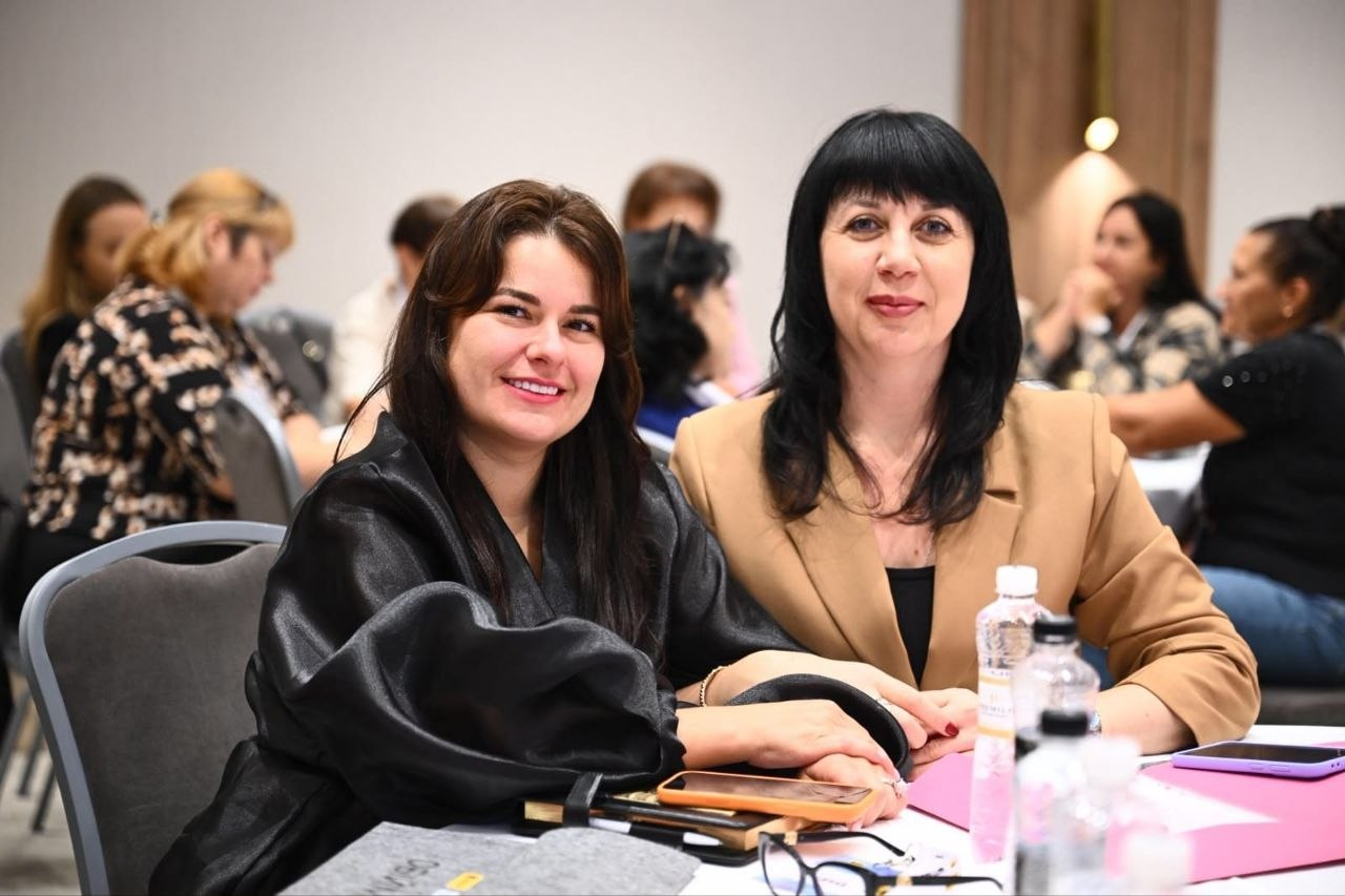 Начальниця служби у справах дітей Херсонської ОДА Тетяна Лушнікова та учасниця зустрічі сидять за столом у конференц-залі під час панельної дискусії «Різні громади — спільна мета» в межах проєкту «Обійми 3.0». На передньому плані ліворуч зафіксовано жінку у чорній блузі з об'ємними рукавами, а праворуч від неї — Тетяну Лушнікову з чорним прямим волоссям у бежевому класичному піджаку, яка посміхається в кадр. На столі перед ними розкладено смартфони, окуляри, папки з паперами та стоїть пляшка води. На задньому плані у розмитті видно інших представників громад та благодійної організації «Українська освітня платформа», які за круглими столами обговорюють механізми підтримки родин Херсонщини та розвиток сімейних форм виховання в умовах війни.
