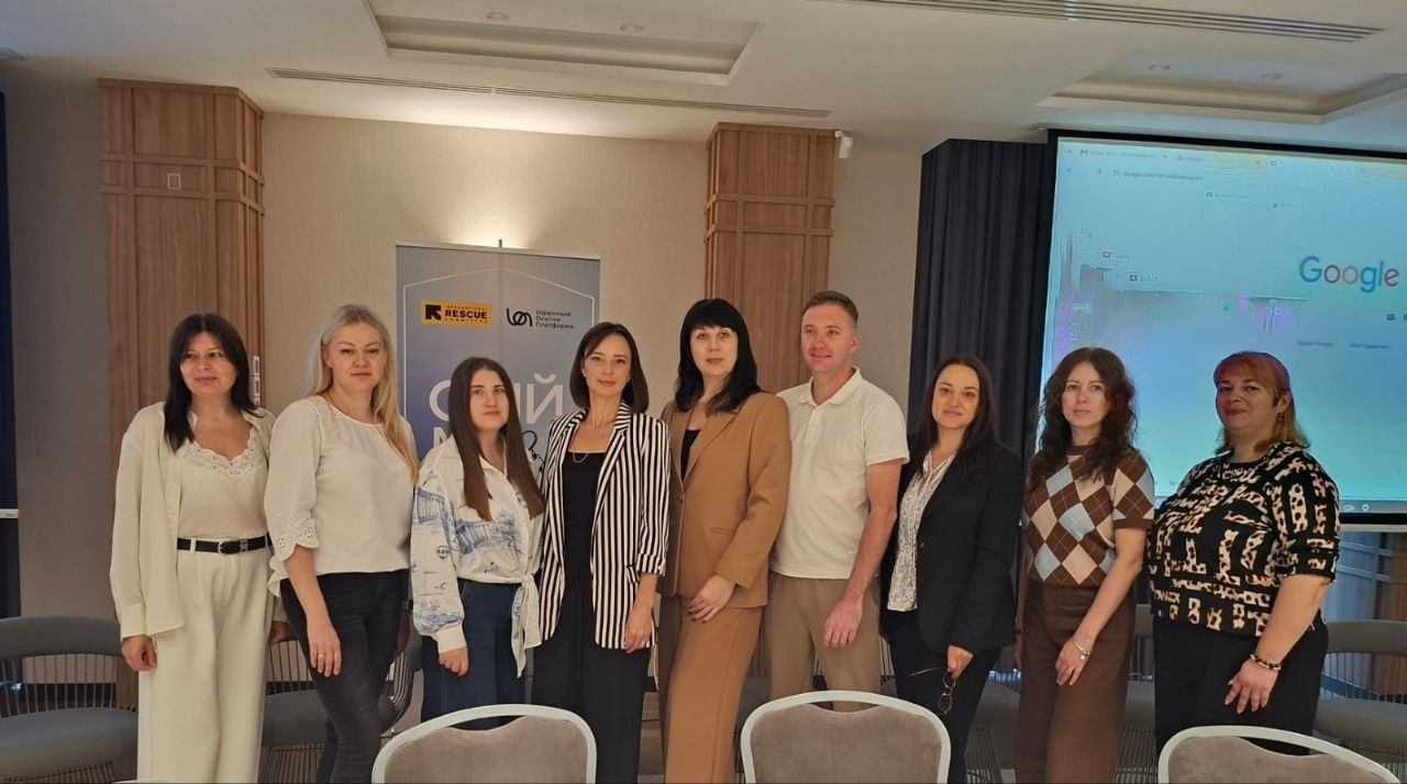 Група учасників та організаторів панельної дискусії «Різні громади — спільна мета» в межах проєкту «Обійми 3.0» позує для спільного фото у світлому конференц-залі. Дев'ятеро людей — вісім жінок та один чоловік — вишикувалися в один ряд по центру приміщення, посміхаючись у кадр. По центру зафіксовано начальницю служби у справах дітей Херсонської ОДА Тетяну Лушнікову в бежевому класичному піджаку та брюках, яка разом із колегами, представниками громад та БО «Українська освітня платформа» обговорила забезпечення прав дитини й підтримку родин в умовах війни. На задньому плані встановлено сірий інформаційний банер із логотипами організації «Rescue» та БО «Українська освітня платформа», а праворуч на стіні розміщено великий мультимедійний екран зі стартовою сторінкою Google.