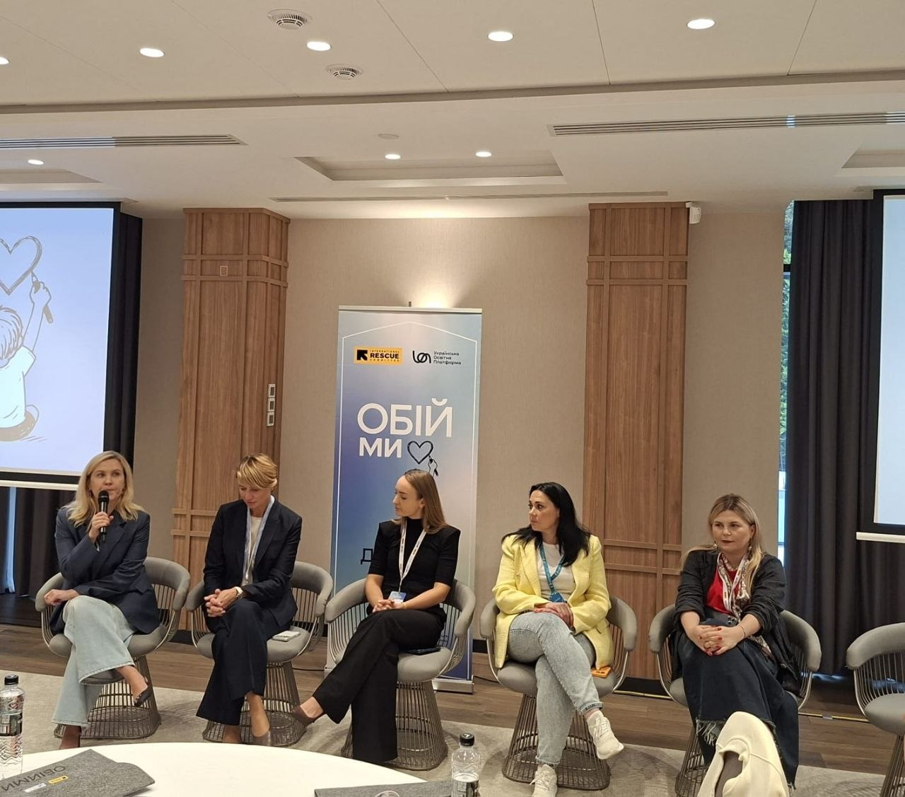 П'ять жінок-спікерок, серед яких начальниця служби у справах дітей Херсонської ОДА Тетяна Лушнікова, сидять у кріслах під час панельної дискусії «Різні громади — спільна мета» в межах проєкту «Обійми 3.0». Учасниці заходу розмістилися в ряд у сучасних сірих плетених кріслах, обговорюючи забезпечення прав дитини та підтримку родин під час війни. Крайня ліворуч жінка в темно-синьому піджаку виступає з мікрофоном у руці. Друга праворуч Тетяна Лушнікова в яскраво-жовтому піджаці та світлих джинсах уважно слухає колег. На задньому плані по центру встановлено блакитний інформаційний банер із логотипами організації «Rescue», БО «Українська освітня платформа» та великим білим написом «ОБІЙМИ» з малюнком серця, а з боків розташовані мультимедійні екрани.